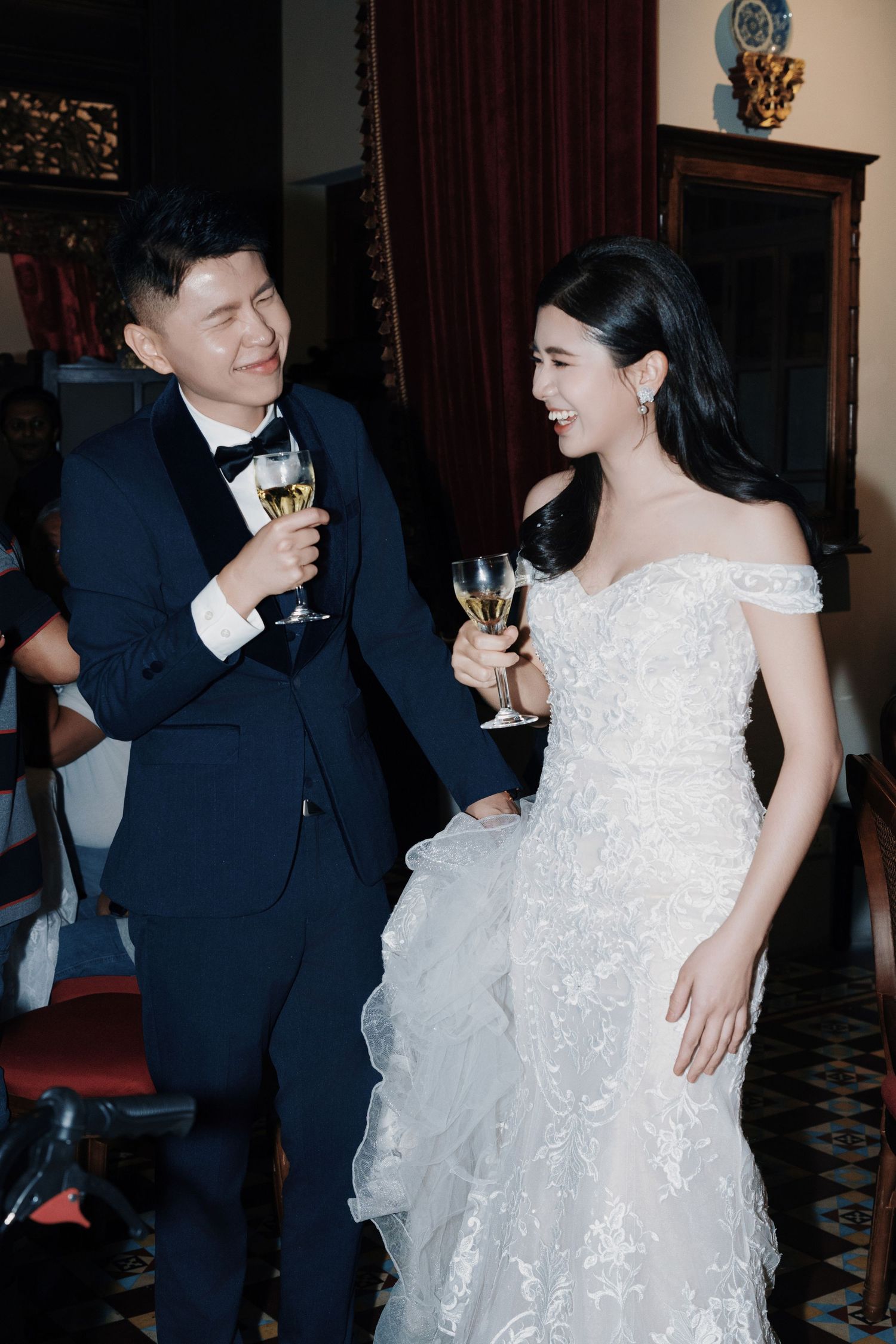 Bride in off-shoulder lace gown and groom in navy tuxedo laughing while holding wine glasses at Kebaya Dining Room