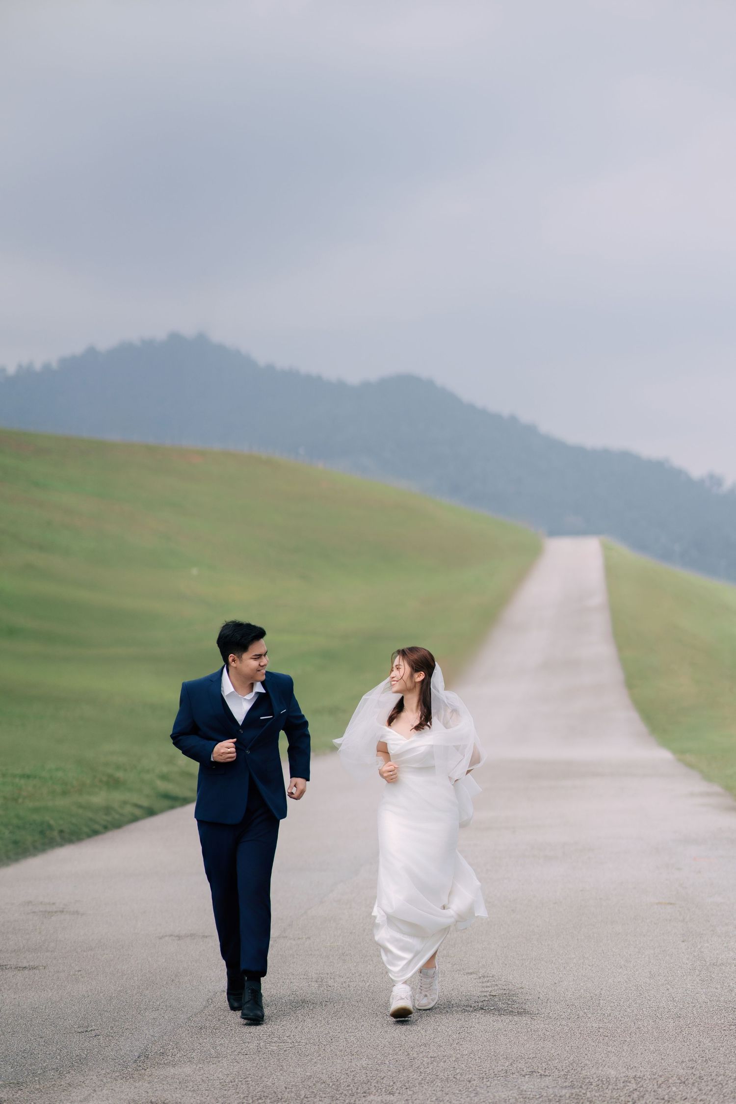 Bride and groom chase each other playfully on an empty mountain road surrounded by green hills.