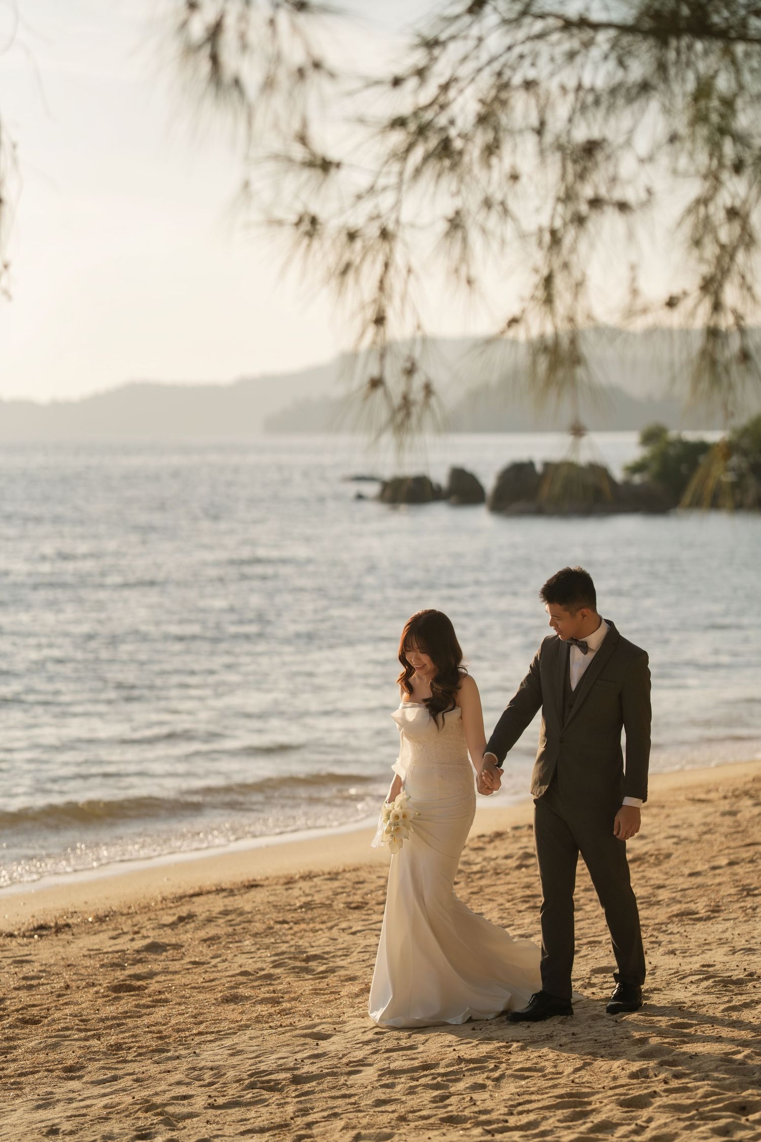 Newlyweds stroll together on a sandy beach with mountains visible in the distance.