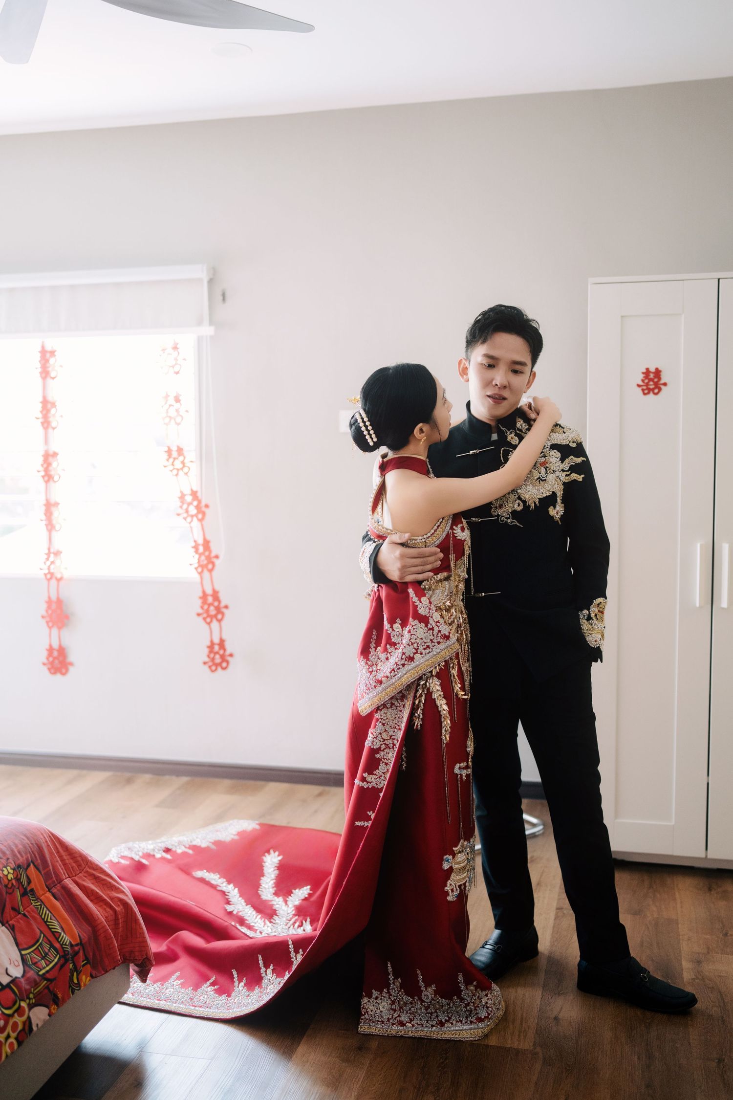 Bride and groom posing in a decorated bedroom wearing traditional Chinese wedding outfits with a red 
