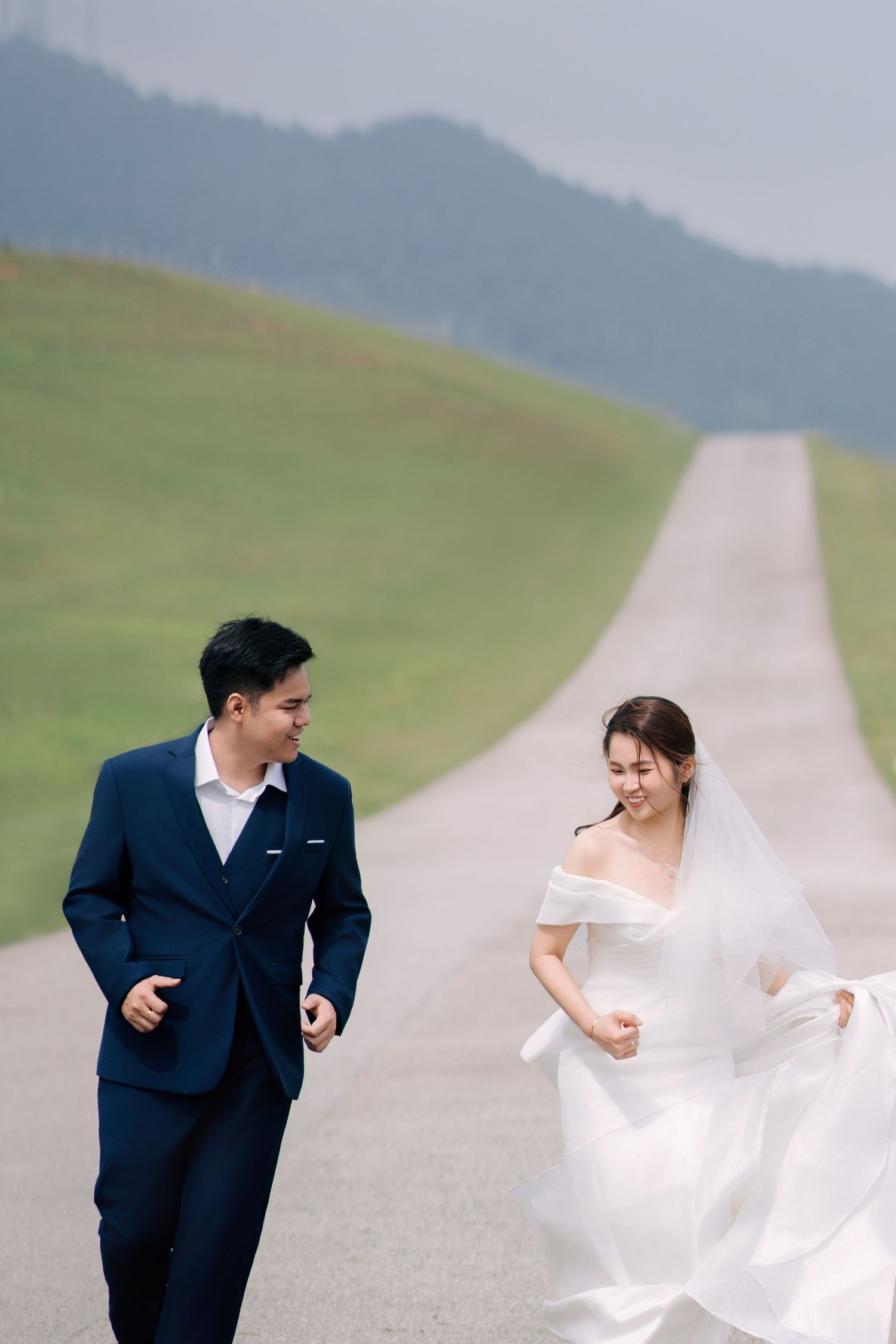 Playful wedding couple running and laughing on a mountain road with rolling green hills in the background.
