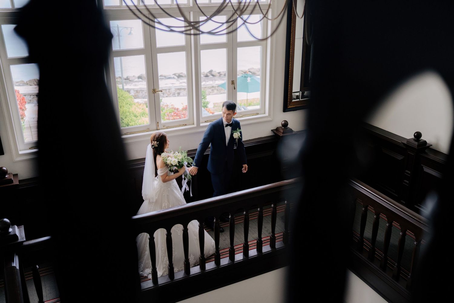A couple descends an elegant staircase with dark wooden railings and large windows in the background at Eastern & Oriental Hotel (E&O).