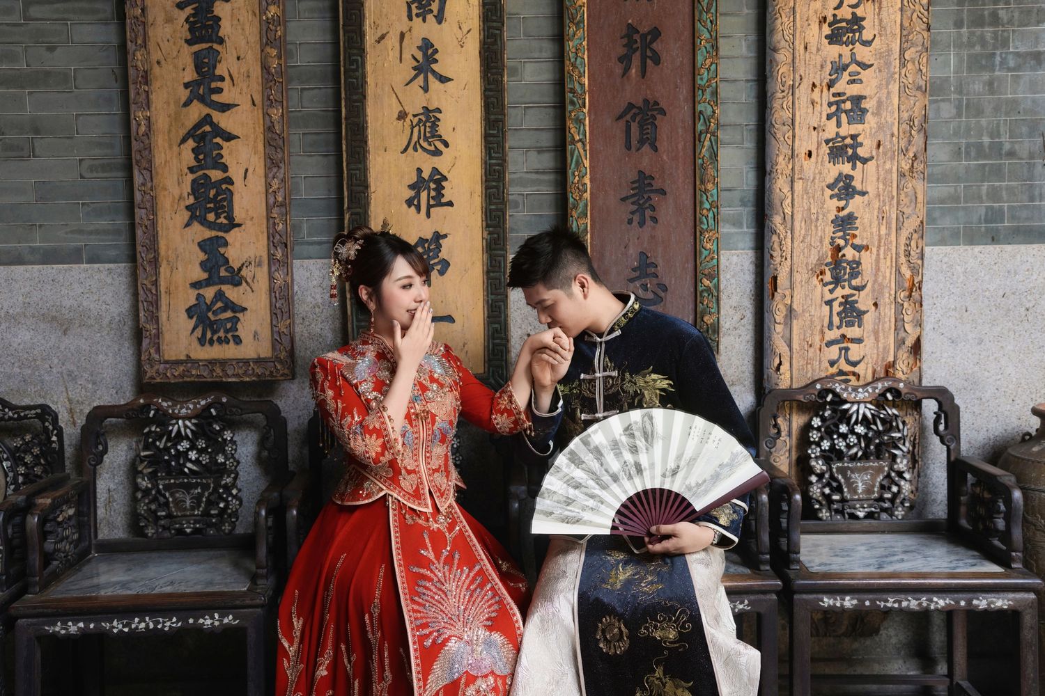 Elegant couple posing in traditional Chinese wedding attire with a paper fan and antique decor