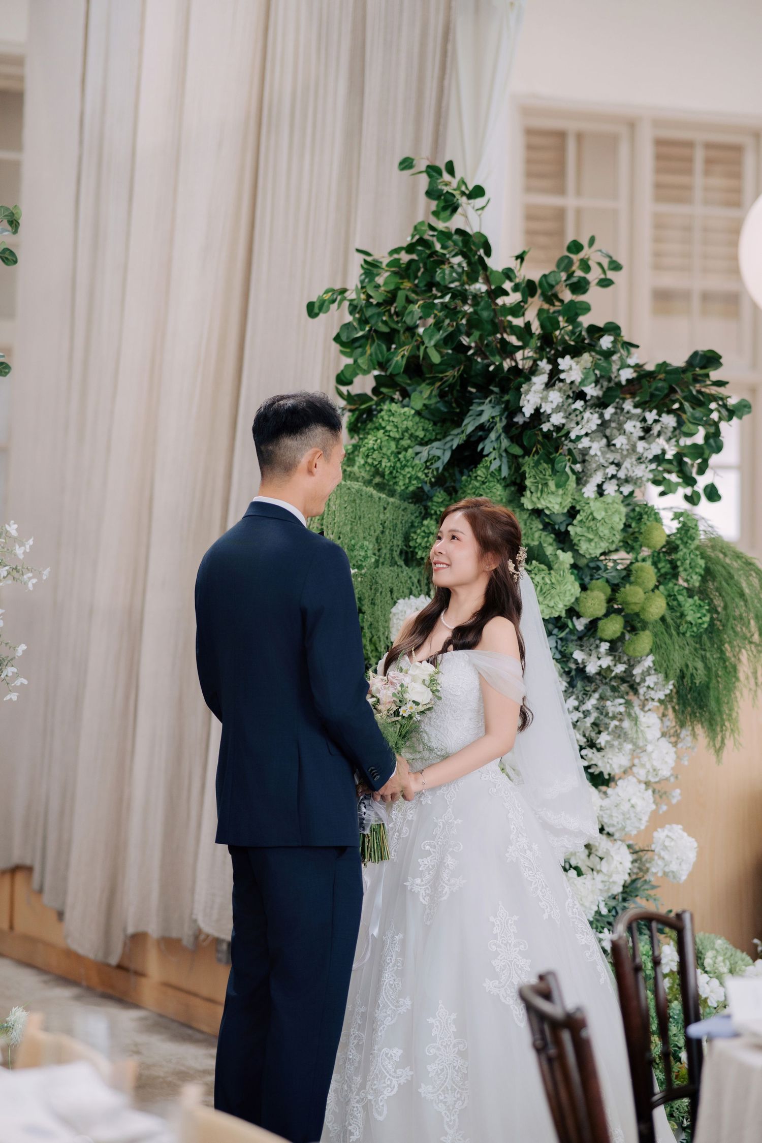 Bride in off-shoulder lace gown and groom in navy suit standing before a tall floral installation
