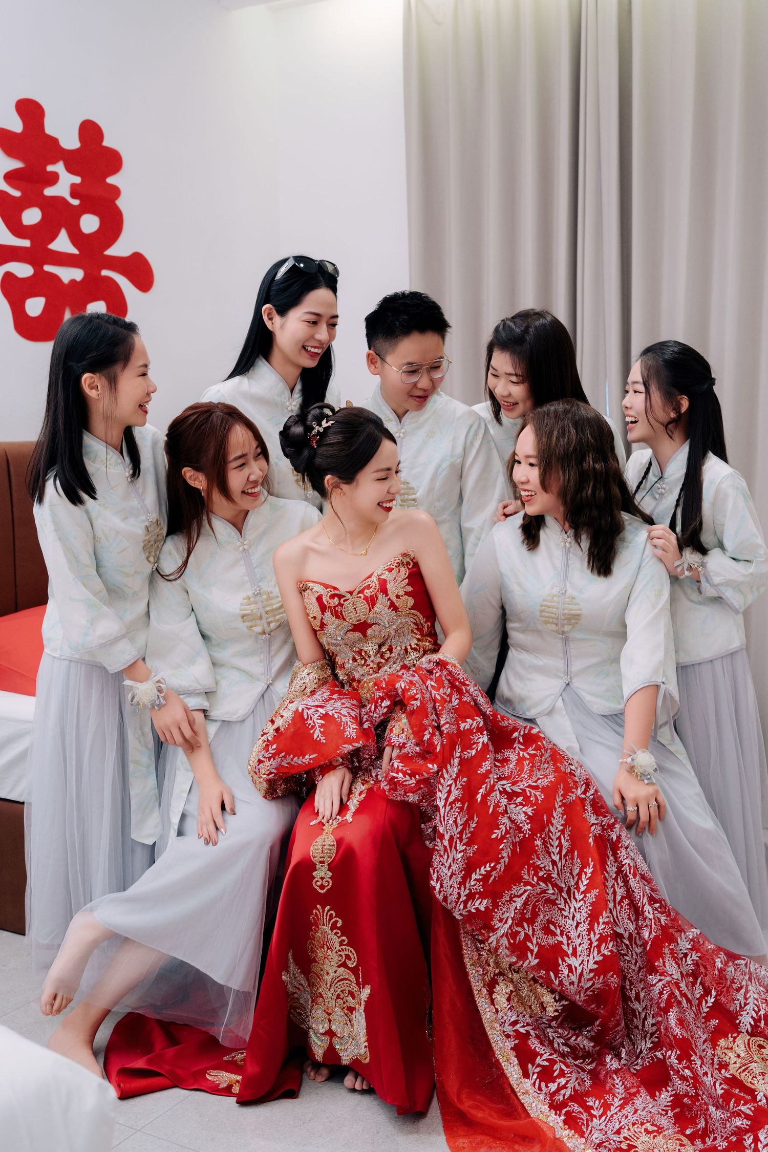 Wedding party members in white outfits gather around someone wearing an ornate red and gold traditional dress.