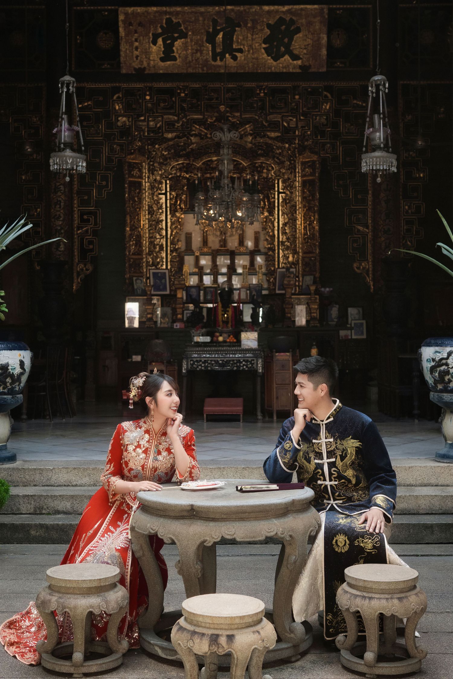 Wedding couple in traditional Chinese outfits seated at stone tables in temple setting.