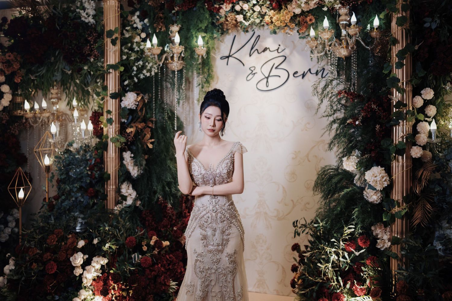 A bride in a heavily beaded champagne gown poses in front of a lush floral wedding backdrop