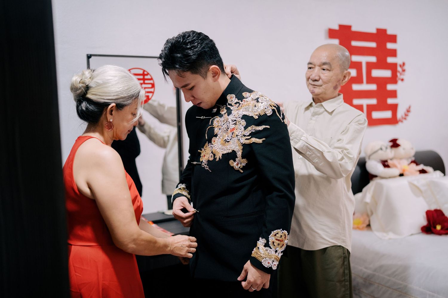 Close-up of parents fastening the buttons on their son's black wedding jacket featuring a gold dragon embroidery