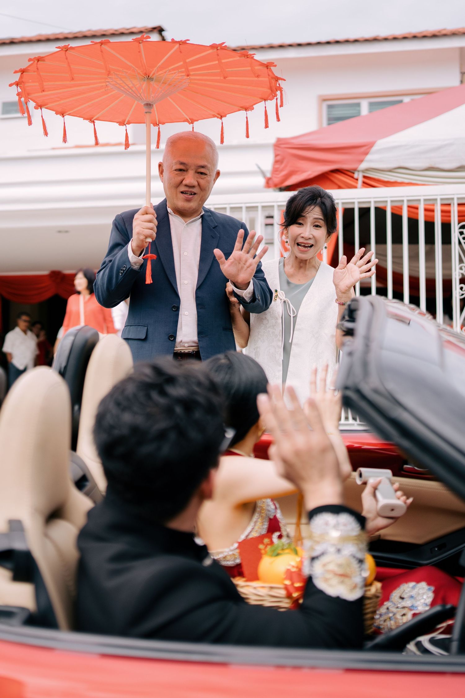 Parents waving under a red umbrella while the bride holds a basket of oranges in a convertible