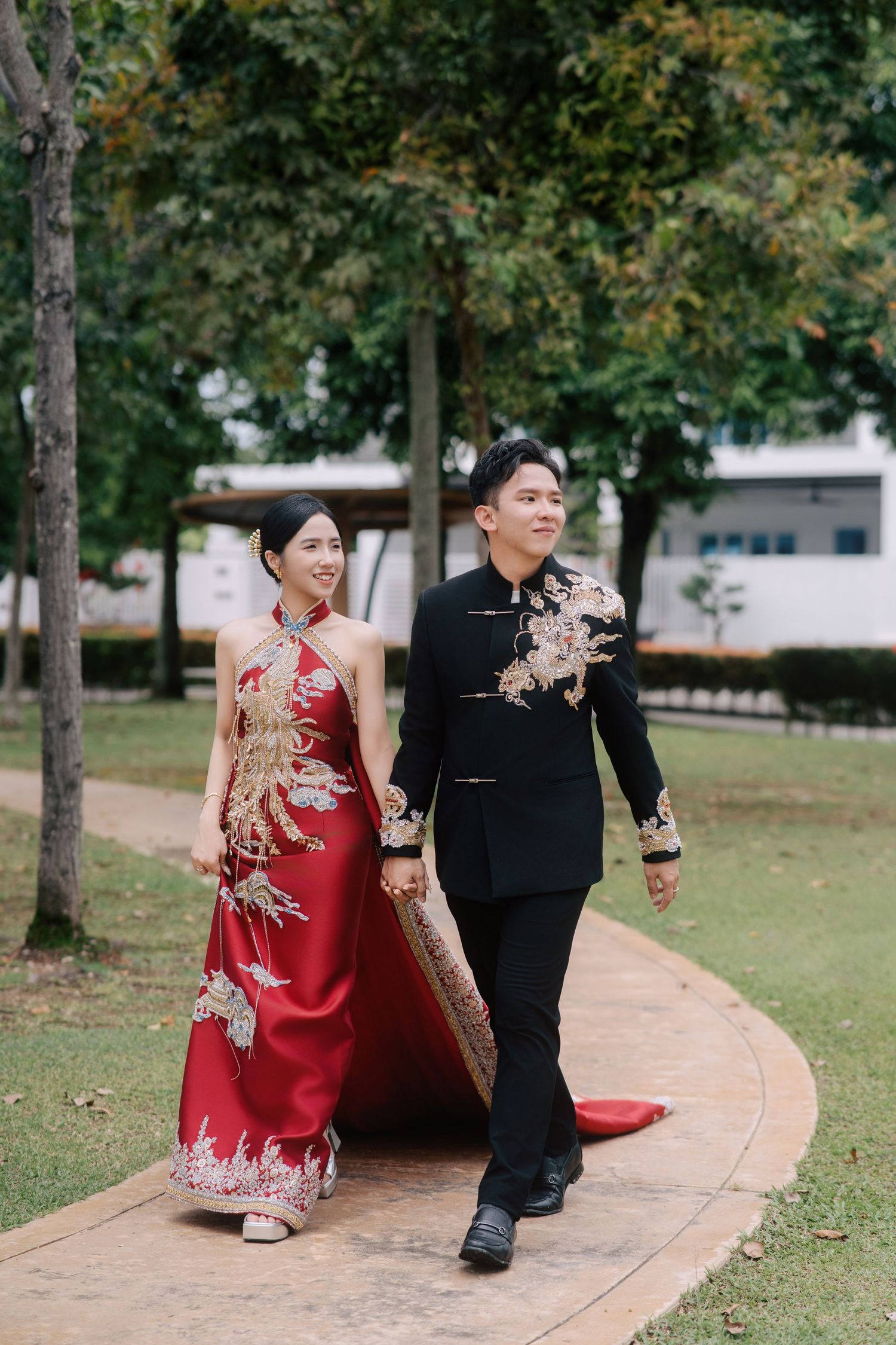 Bride and groom in custom traditional Chinese wedding attire walking outside, featuring a red phoenix qipao and black dragon suit