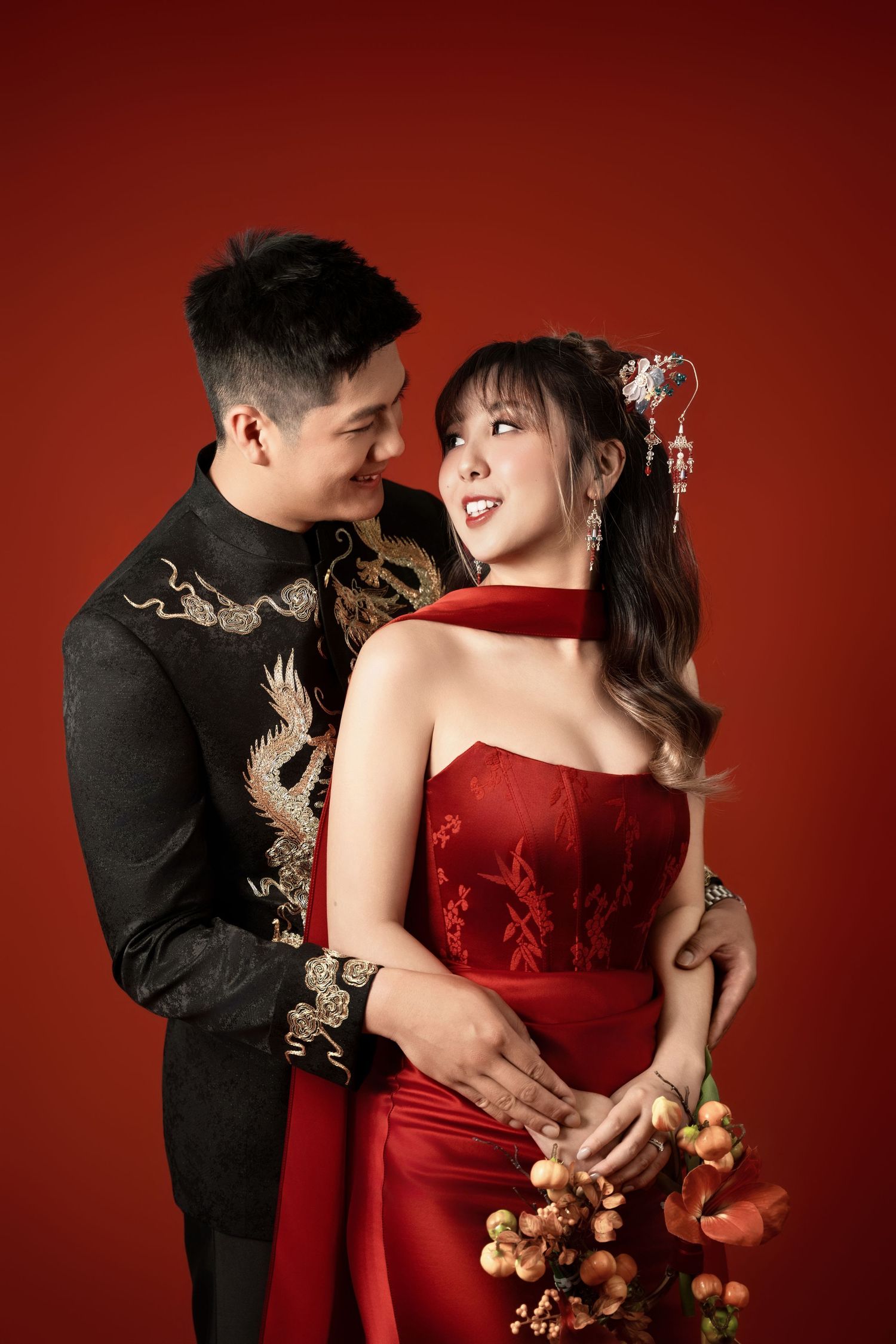 Romantic couple poses in red dress and black dragon-embroidered suit against deep red backdrop.