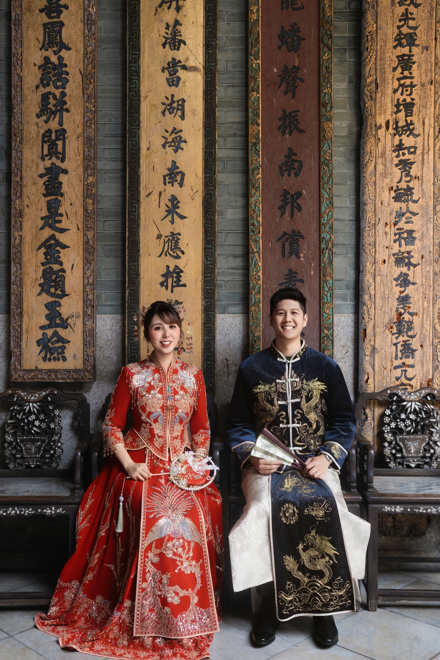 Traditional Chinese wedding photos with couple in red qipao and black embroidered suit against calligraphy wall.