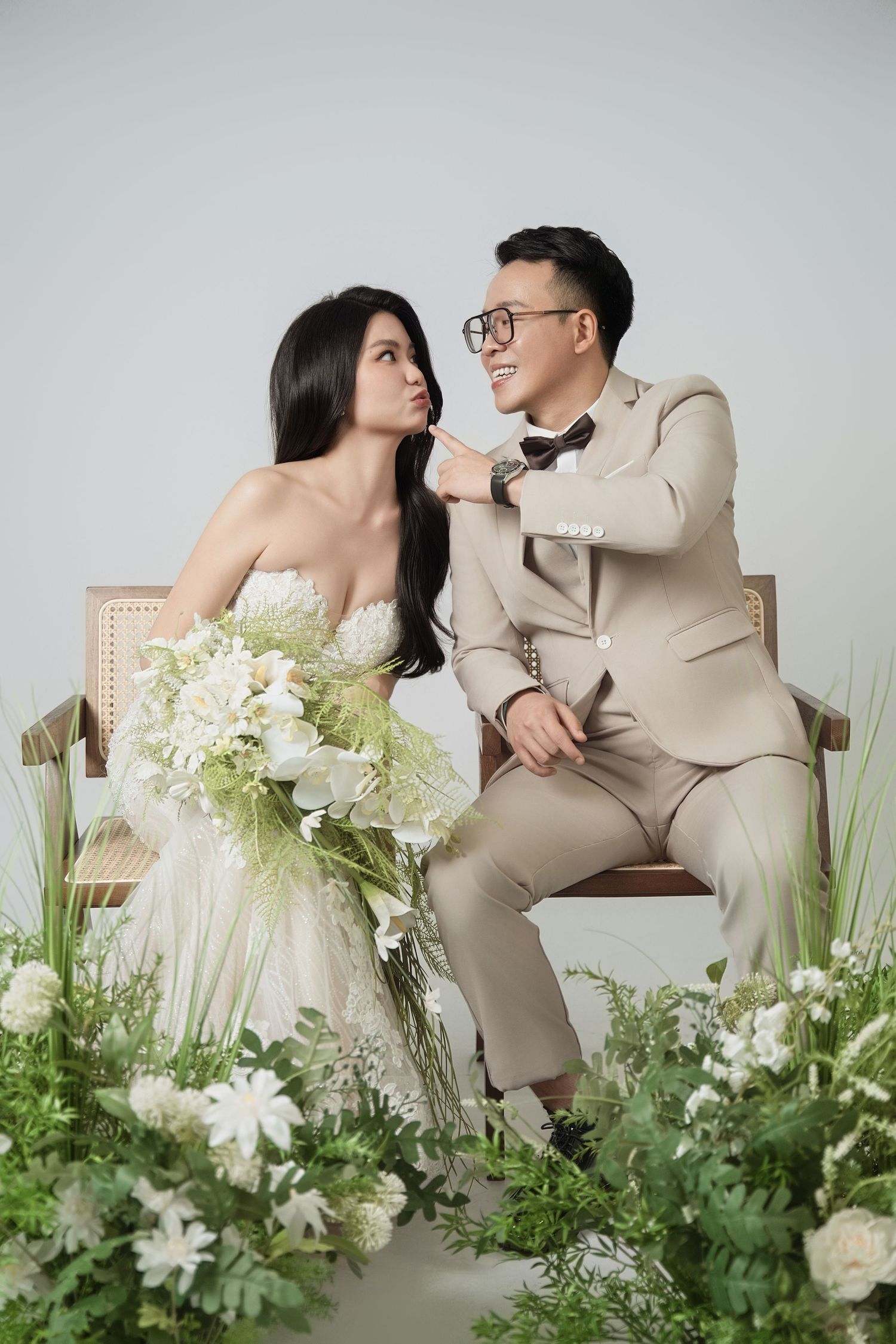 Wedding couple poses playfully on wooden chairs surrounded by white and green floral arrangements.
