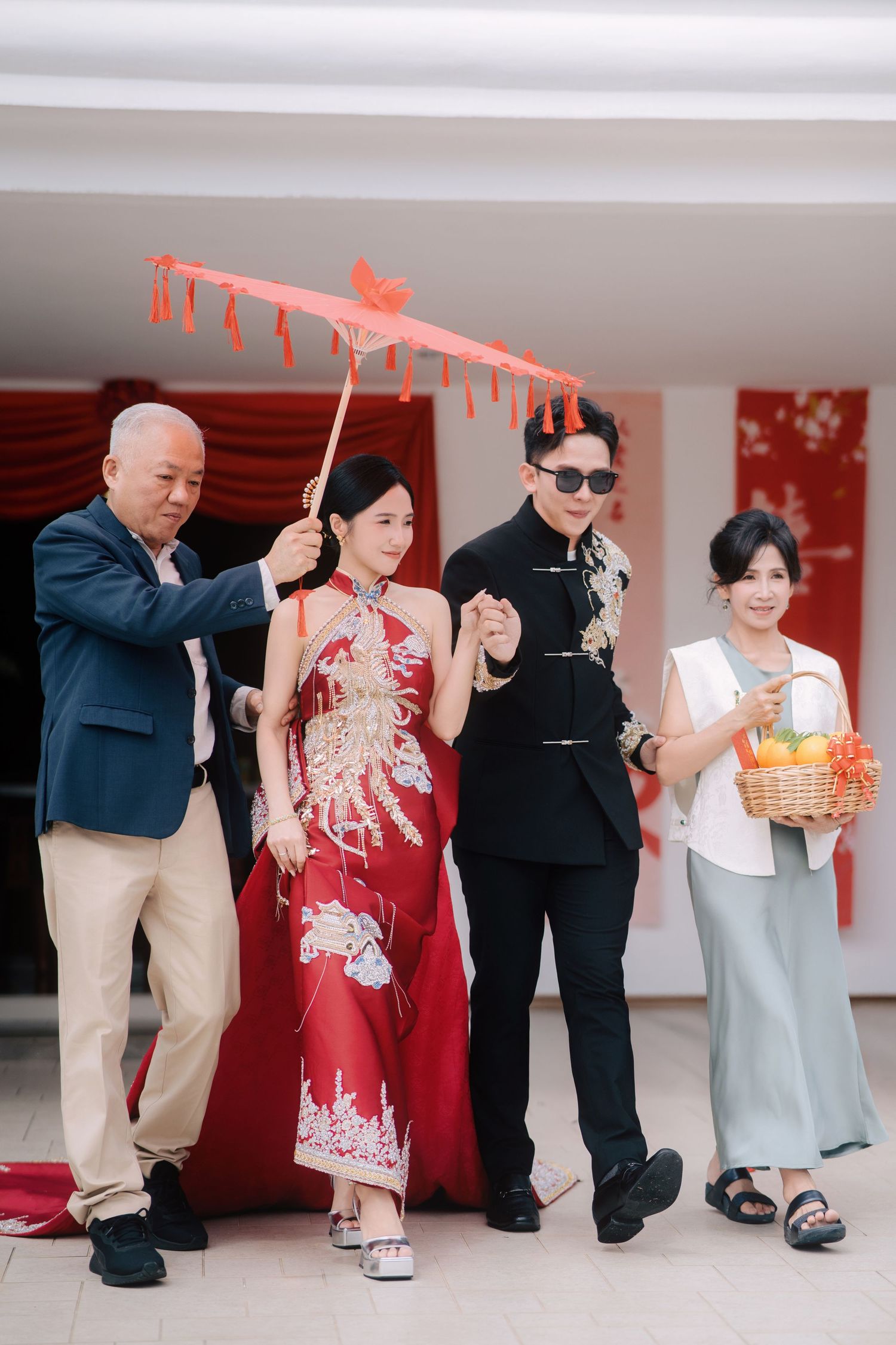 The bride’s father holds a red oil-paper umbrella over the couple as they walk outside, accompanied by the mother