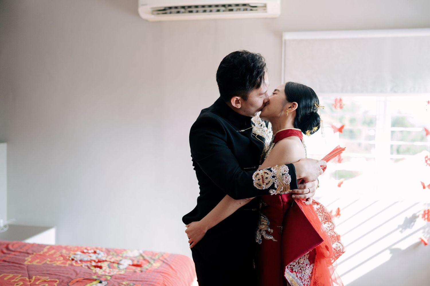 Newlywed couple in traditional red and black Chinese attire sharing a kiss in a decorated bedroom