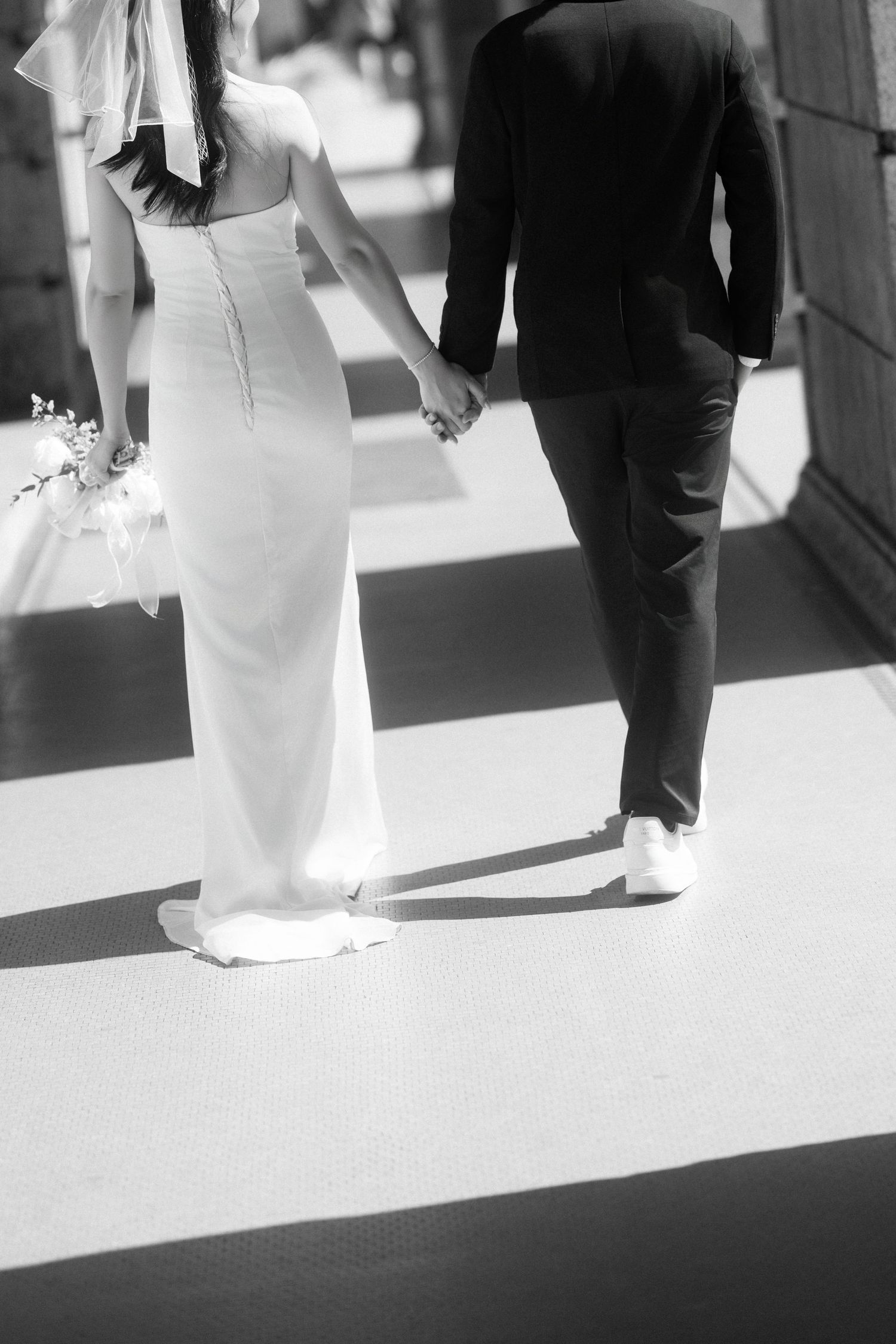 Black and white photo of a couple walking hand in hand, showing elegant wedding attire from behind.