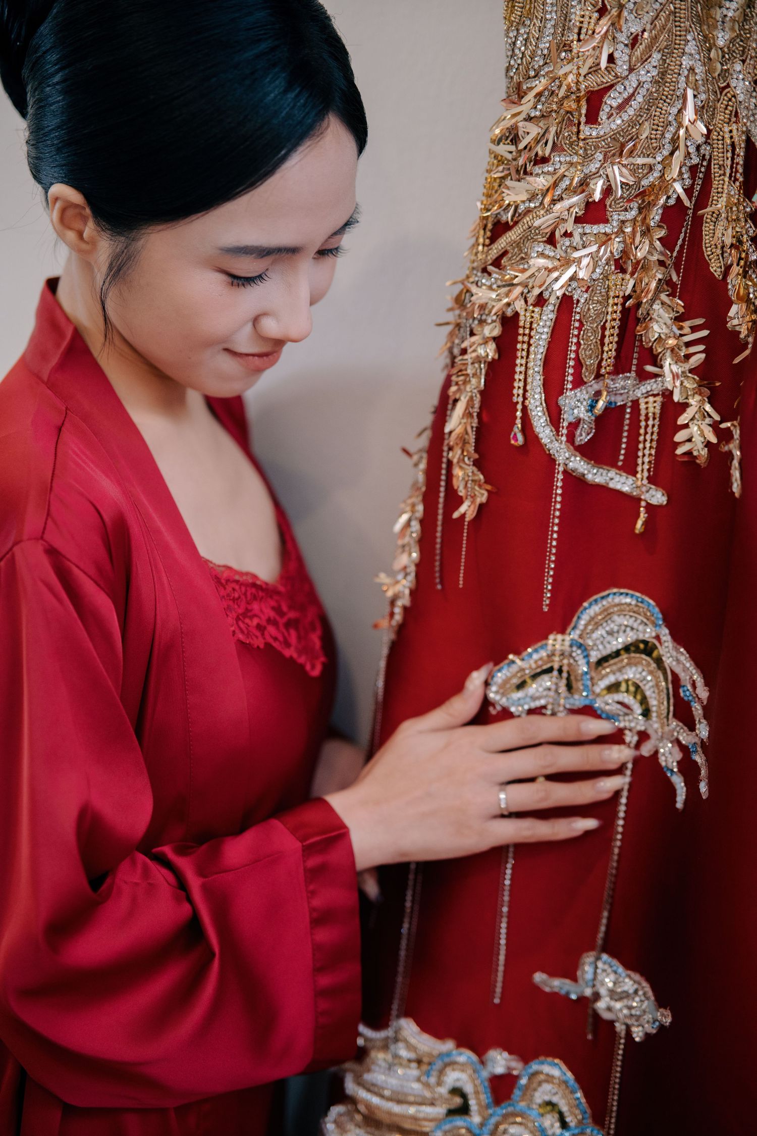 A bride in a red silk robe gently touching the intricate gold embroidery of her traditional wedding gown