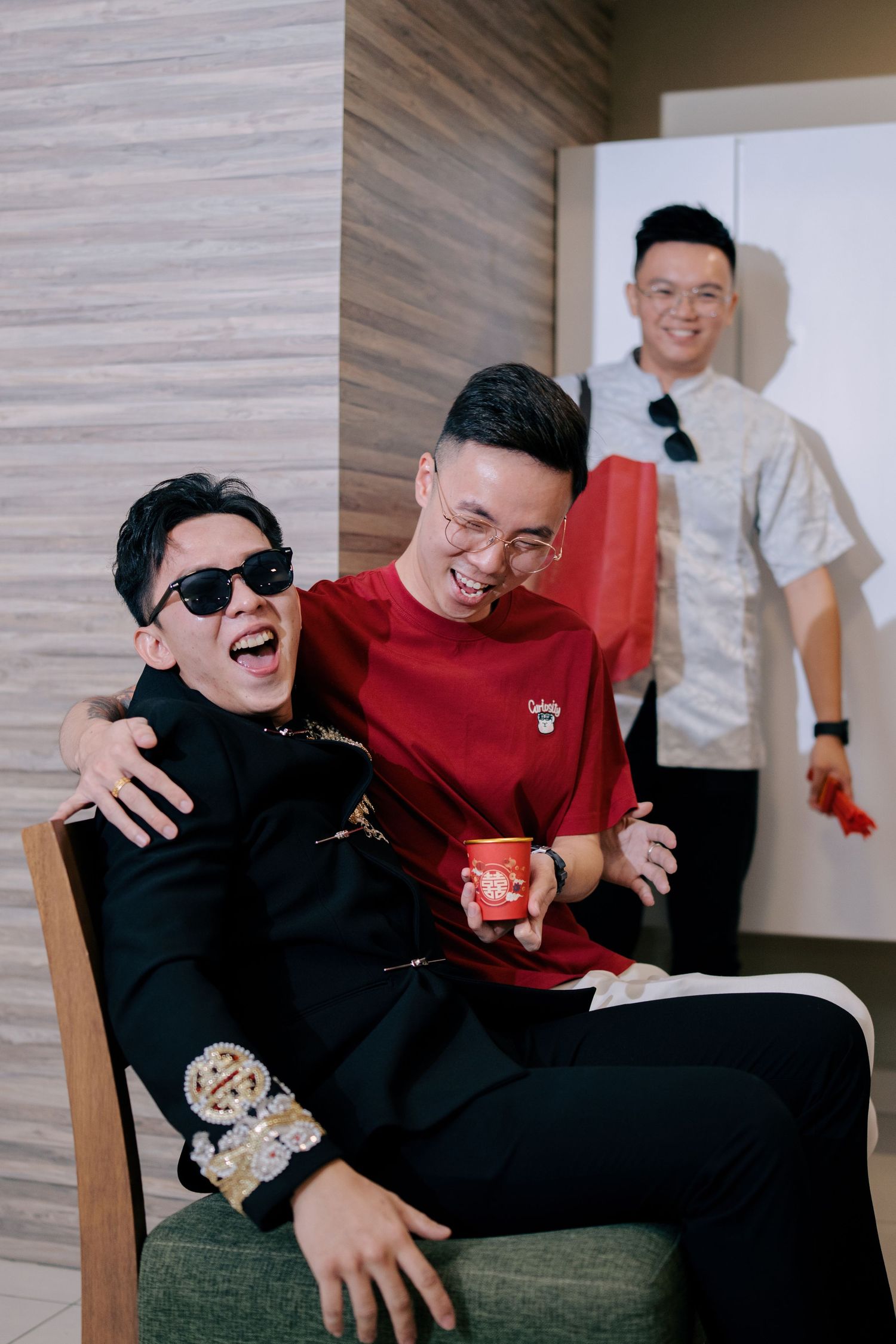Groom laughing in a chair while a friend holds a red cup with a double happiness symbol