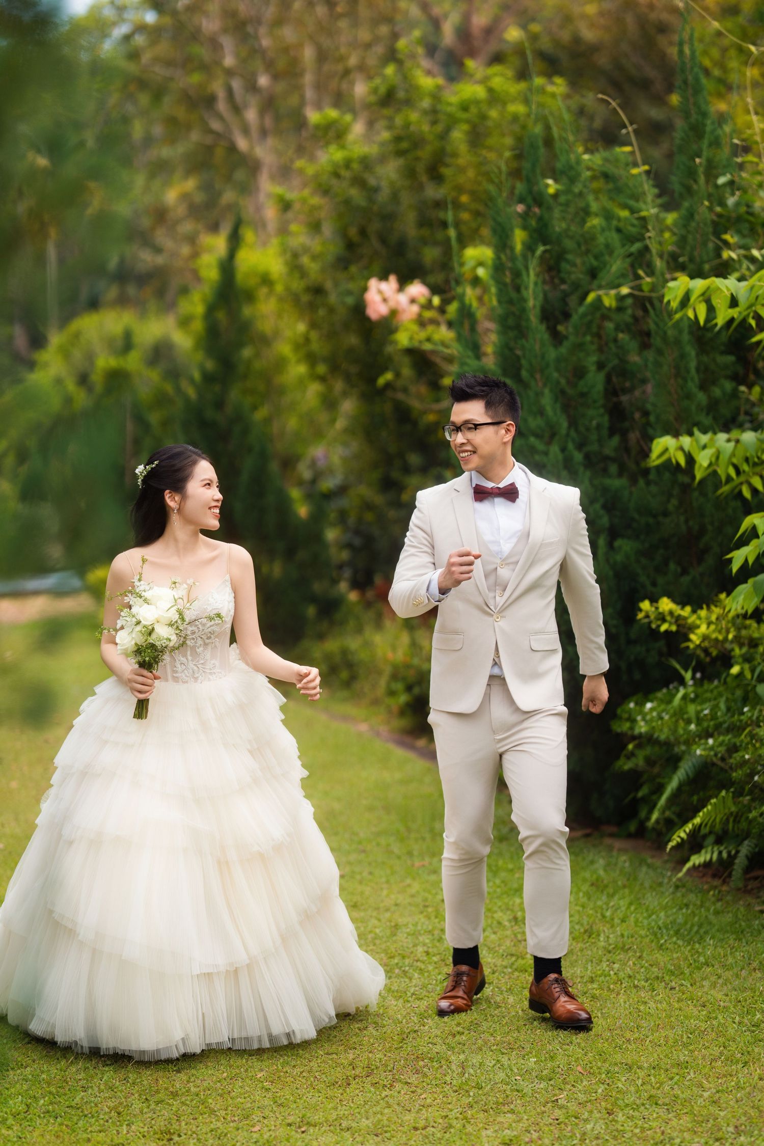 A couple in white wedding attire playfully poses in a lush garden setting.