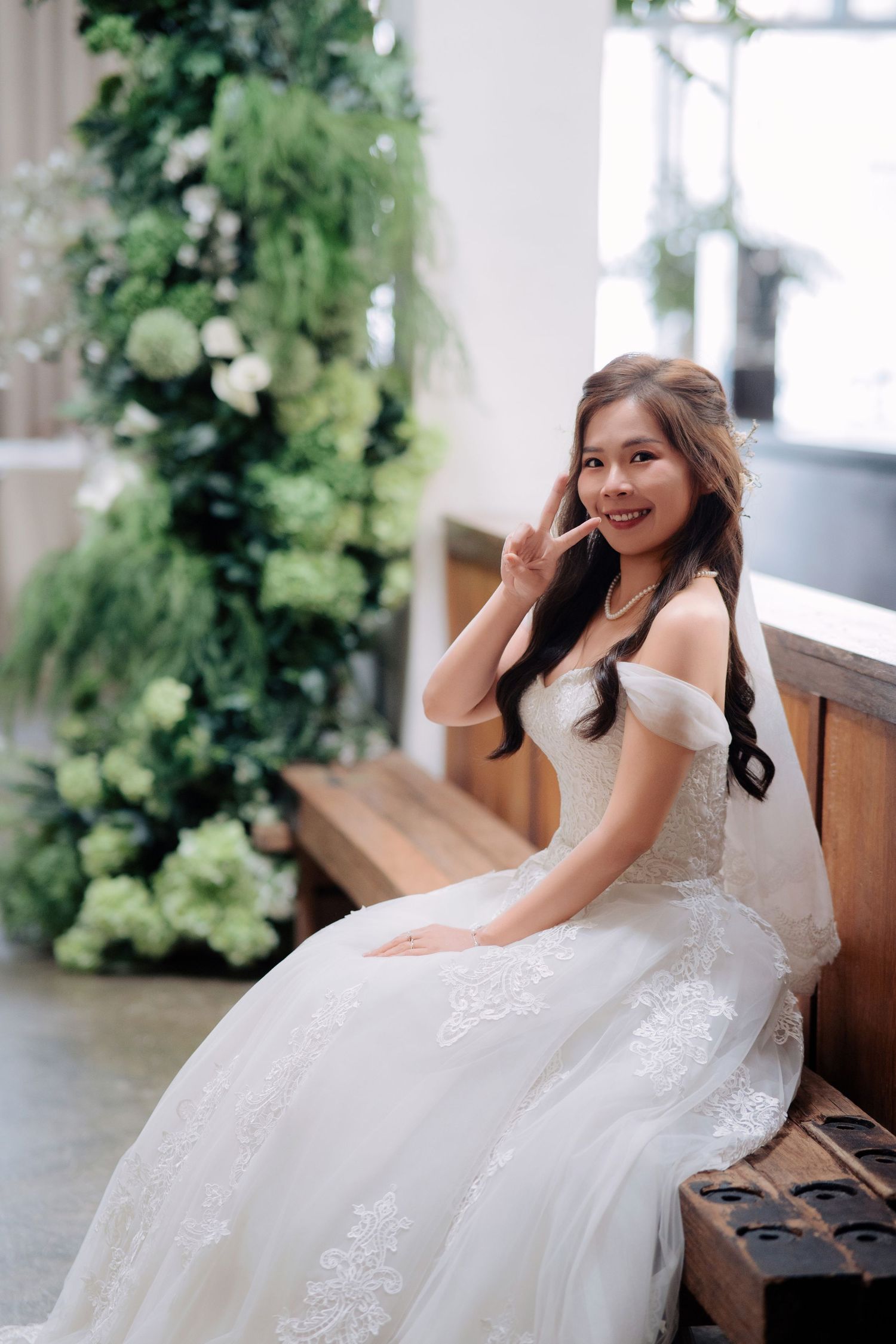 Bride in an off-the-shoulder lace wedding dress sitting on a wooden bench in front of floral pillars