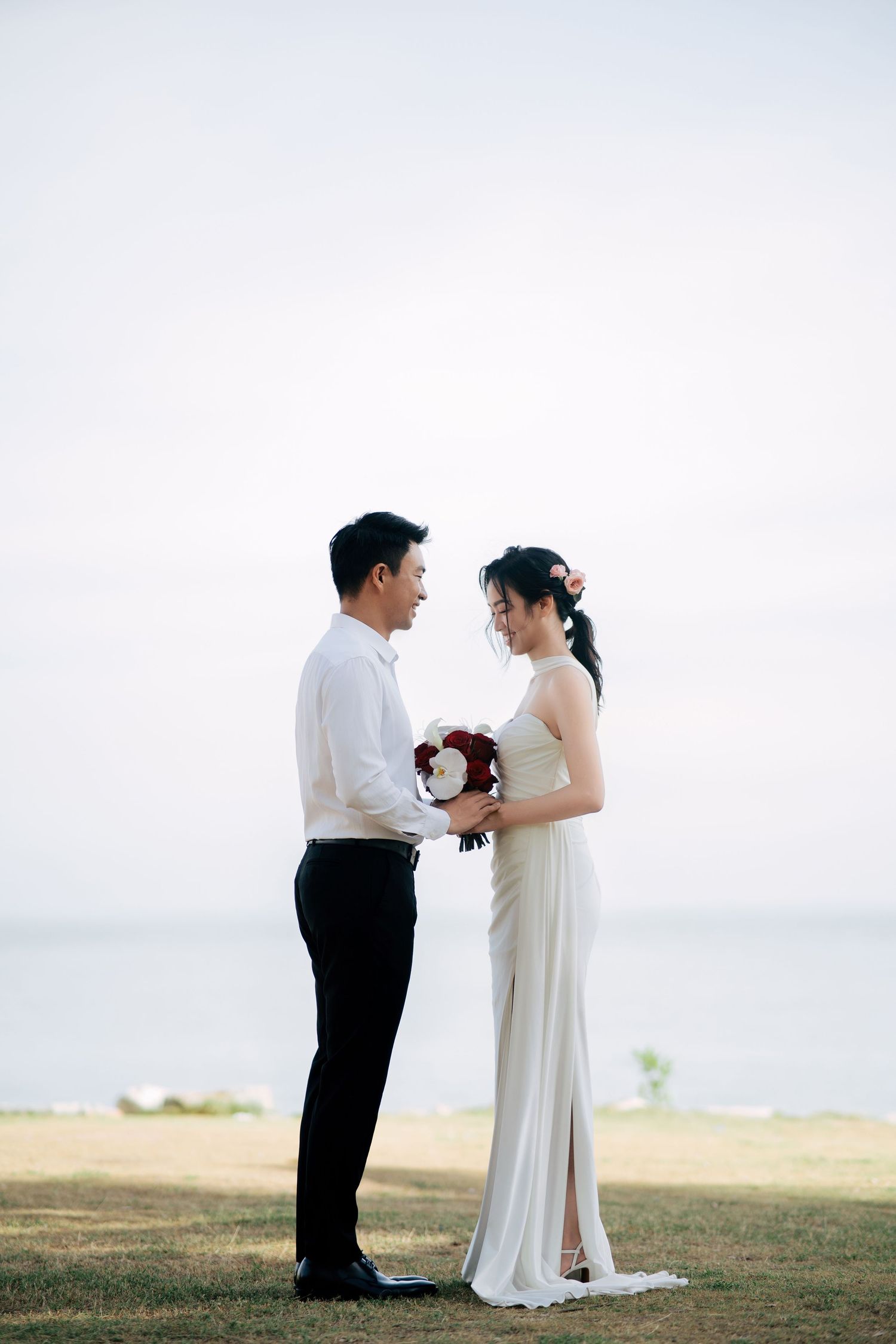 A couple in wedding attire stands close together on a grassy hill overlooking the ocean.