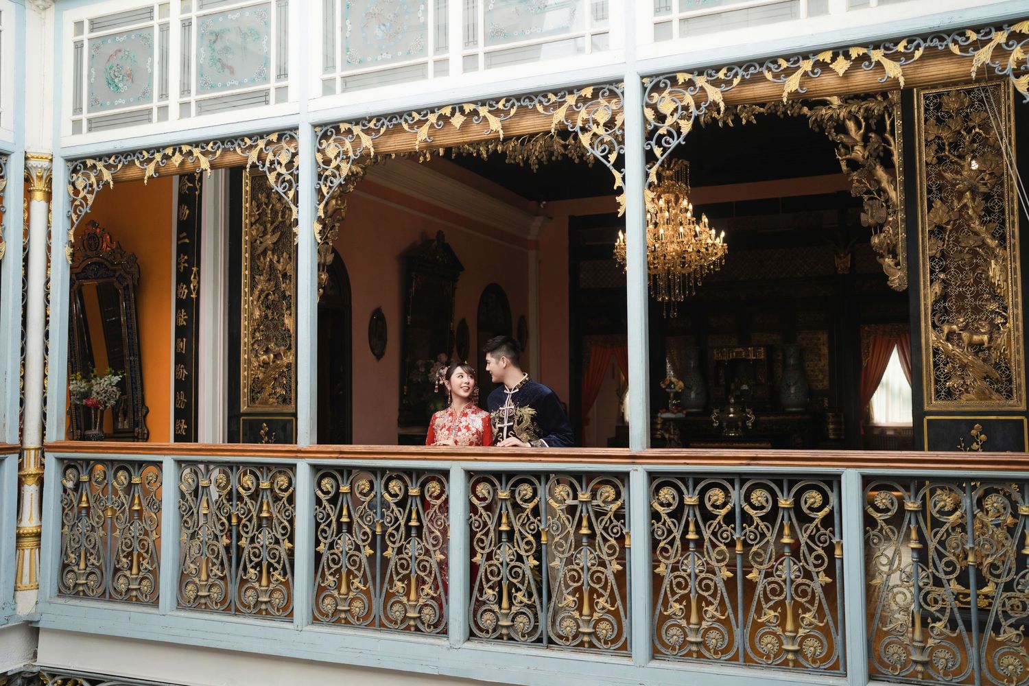 Couple looking at each other while posing on a historic balcony decorated with gold Chinese carvings and blue railings