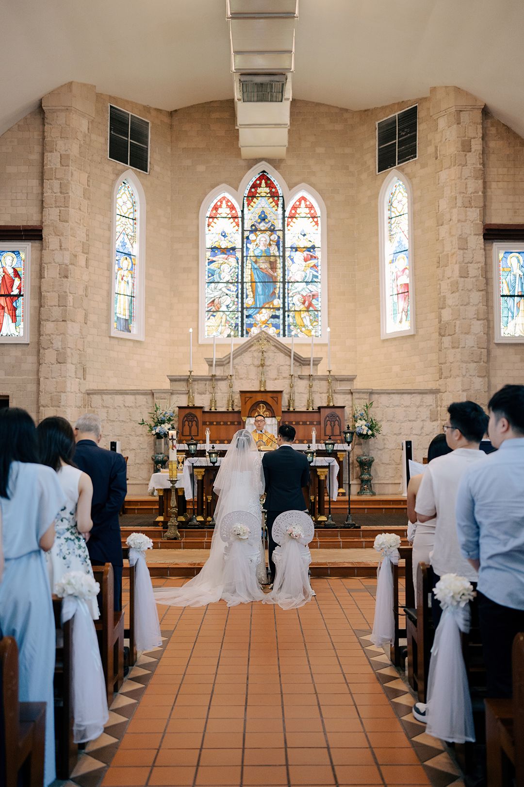 Catholic church wedding ceremony featuring a bride and groom kneeling before a stained-glass altar