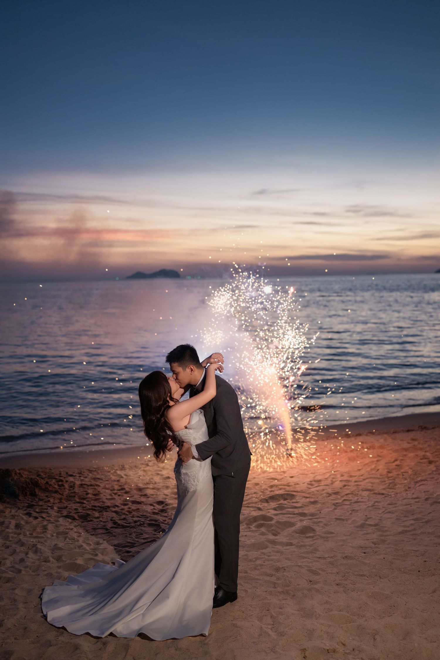 Dramatic beach wedding portrait with sparklers illuminating the twilight sky.