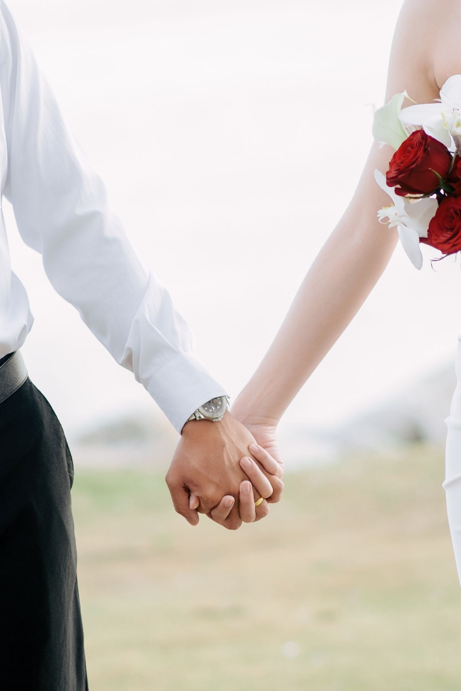 Close-up of a couple holding hands with red roses against a blurred outdoor background.