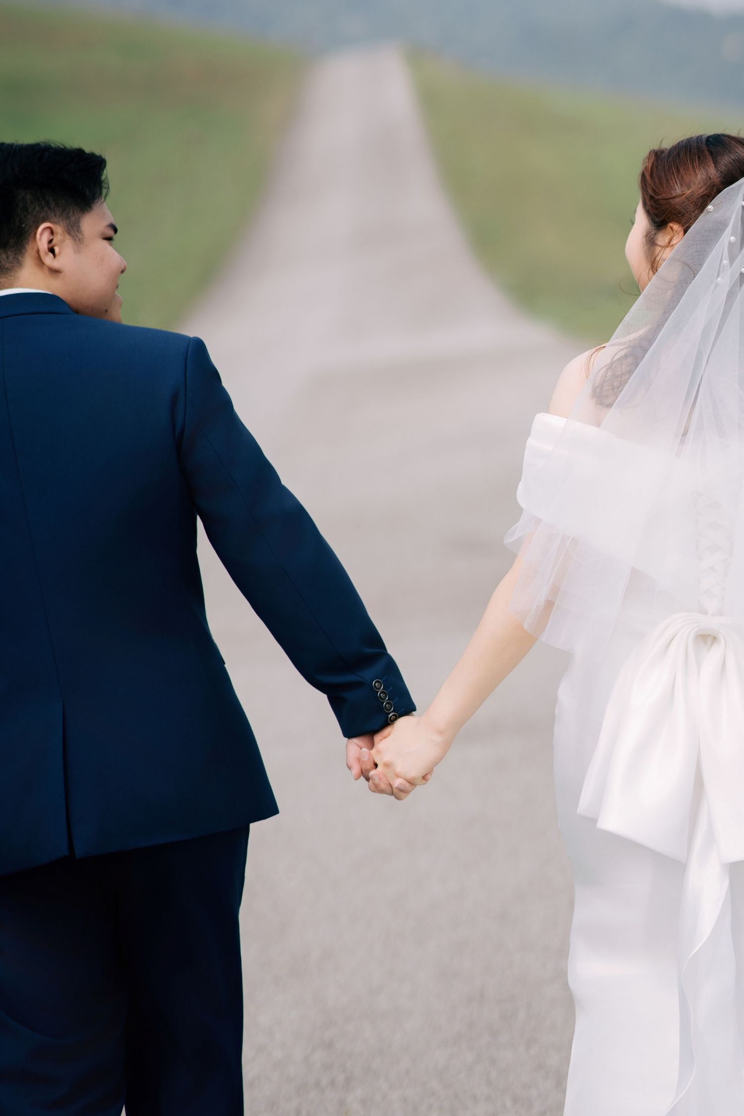 A couple holds hands while walking down a long road through green hills.