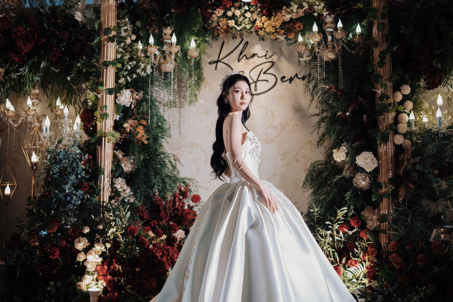 Portrait of a bride in a sparkling white gown standing before a lush, romantic floral wedding stage at Olive Tree Hotel