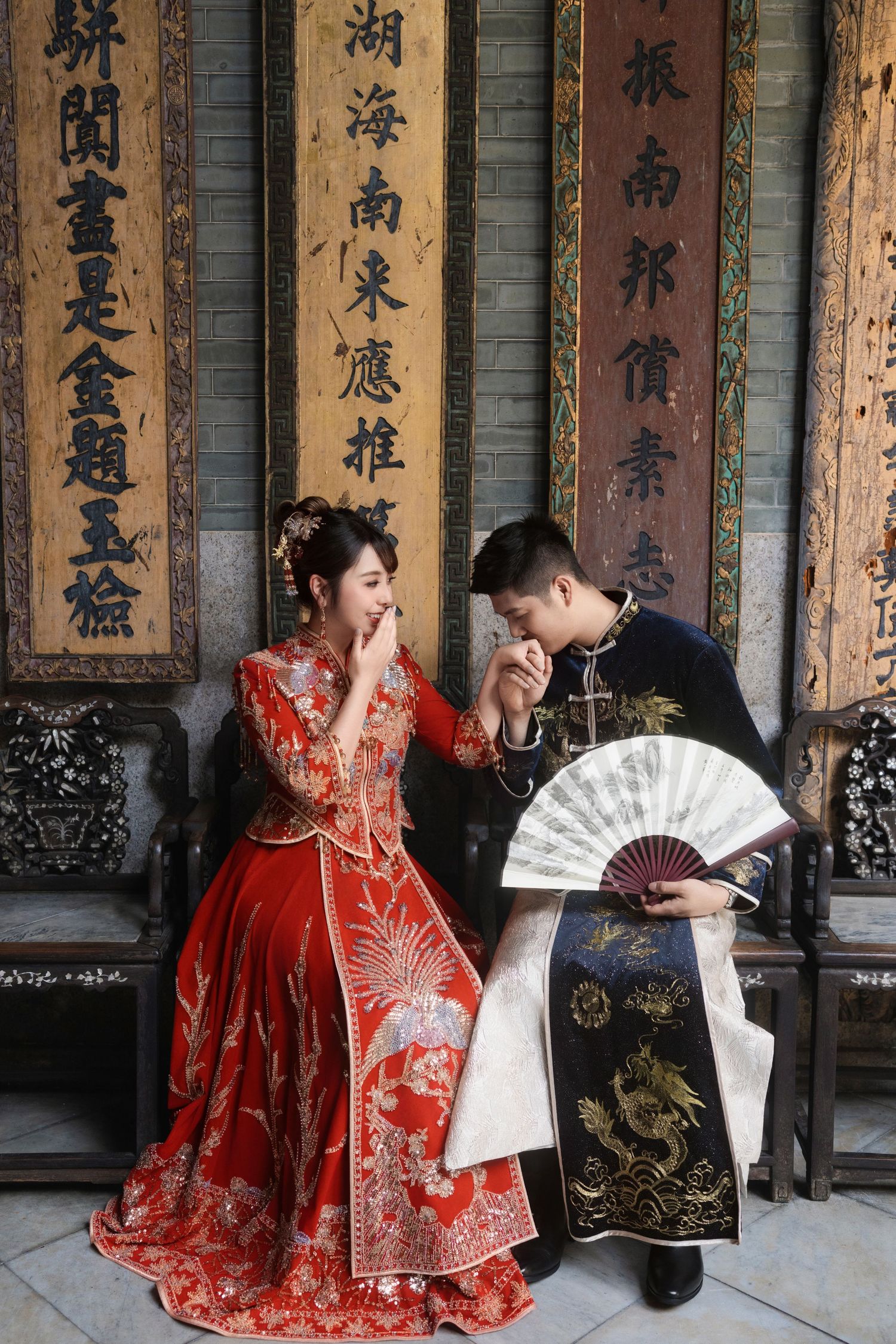 Groom in a navy robe kissing the bride's hand while she laughs, both in traditional Chinese wedding attire