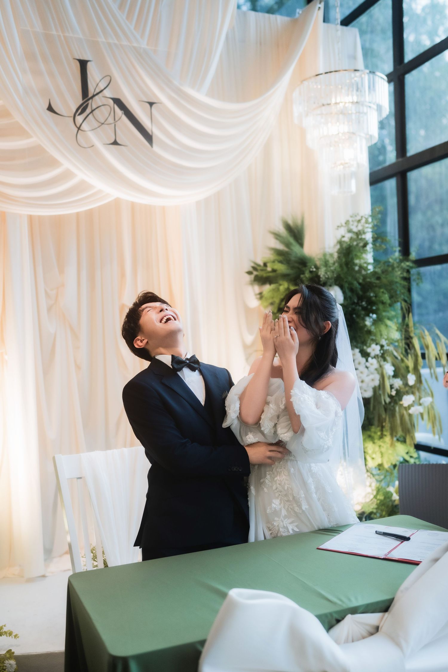 A couple shares a joyful moment signing papers at a wedding ceremony with white drapes.
