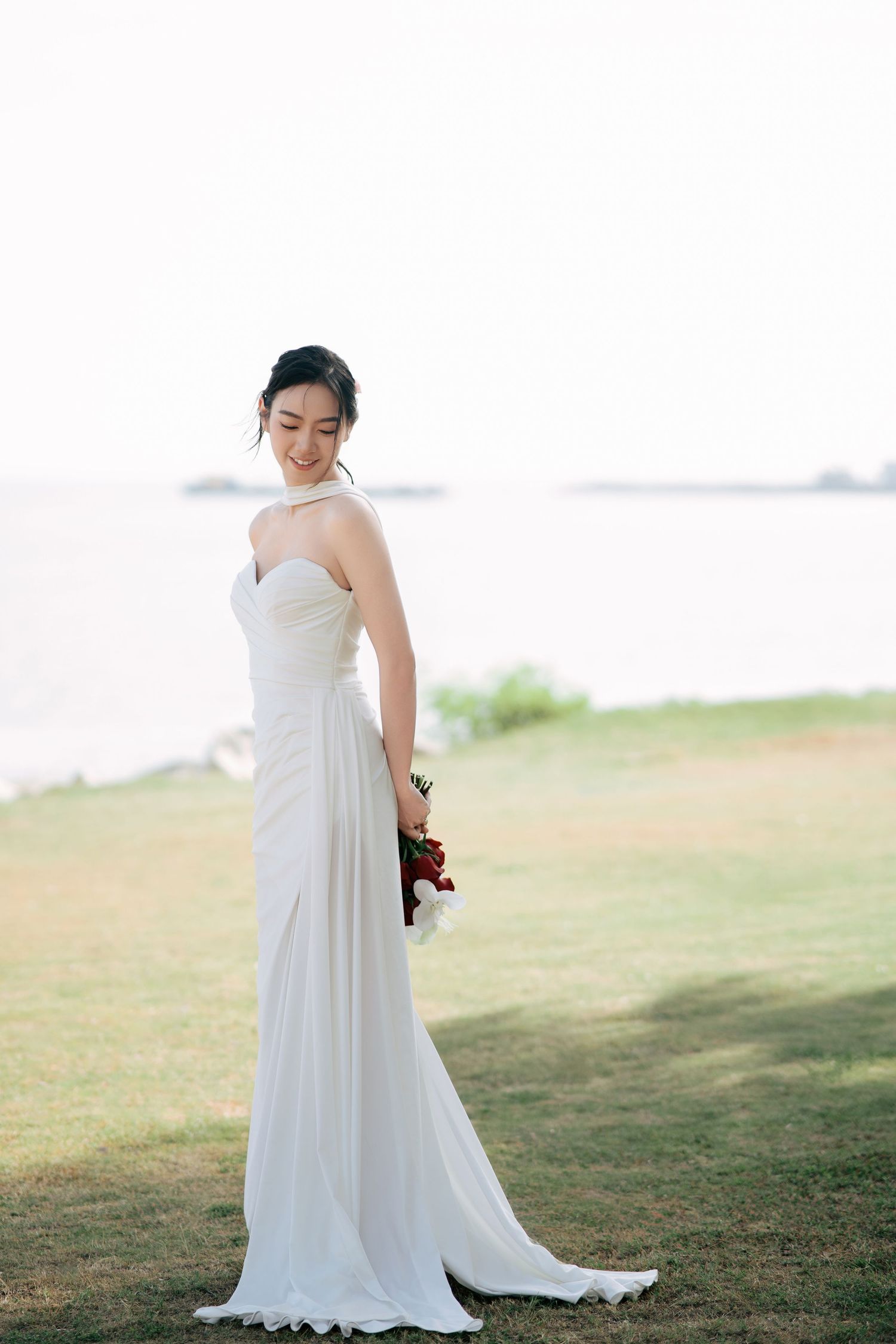 An elegant bride in a flowing white dress poses on a grassy hillside by the water.
