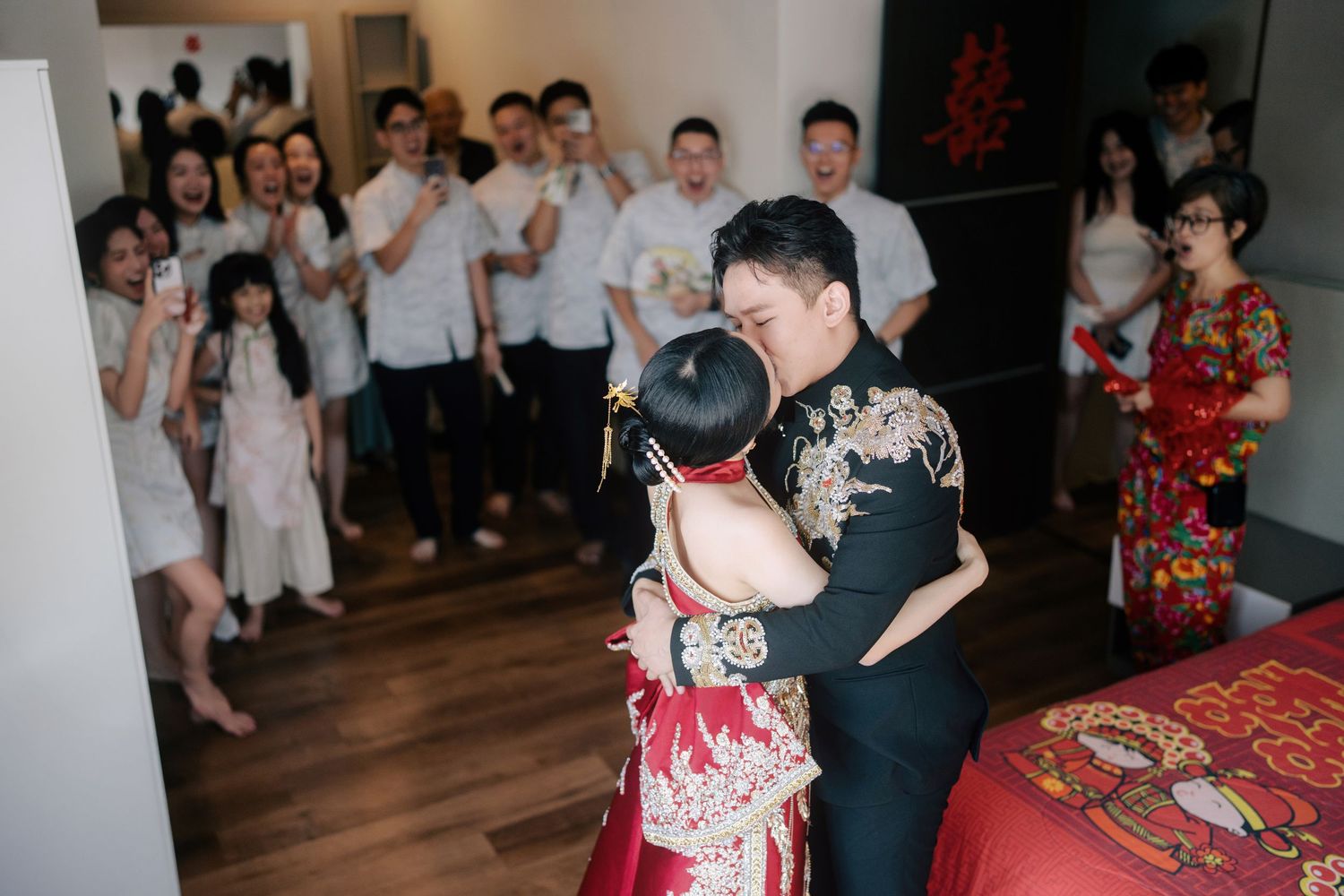 Bride and groom in traditional Chinese wedding attire kissing as bridesmaids and groomsmen cheer in the background