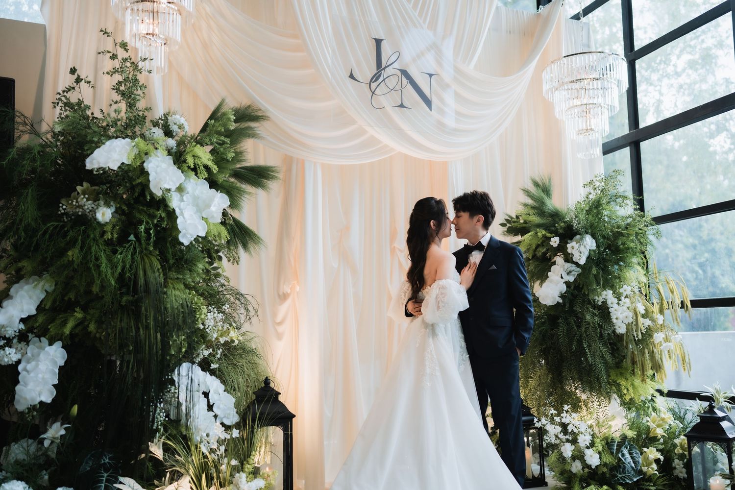 Bride and groom posing by a modern indoor wedding floral backdrop featuring draped white fabric, personalized monograms, and lush greenery