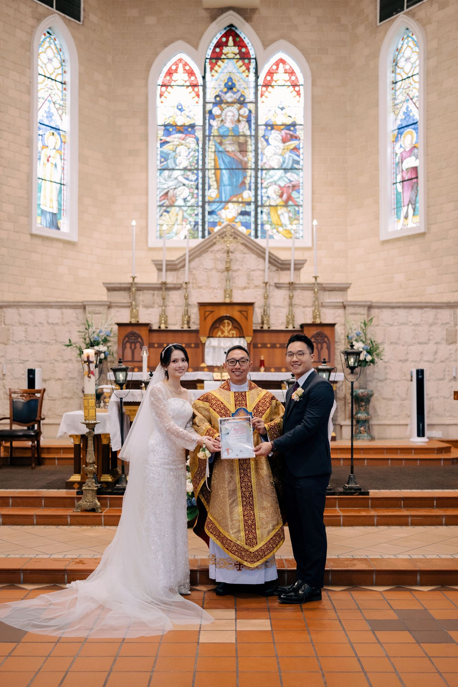 Newlyweds posing with the priest and marriage certificate at a Catholic altar