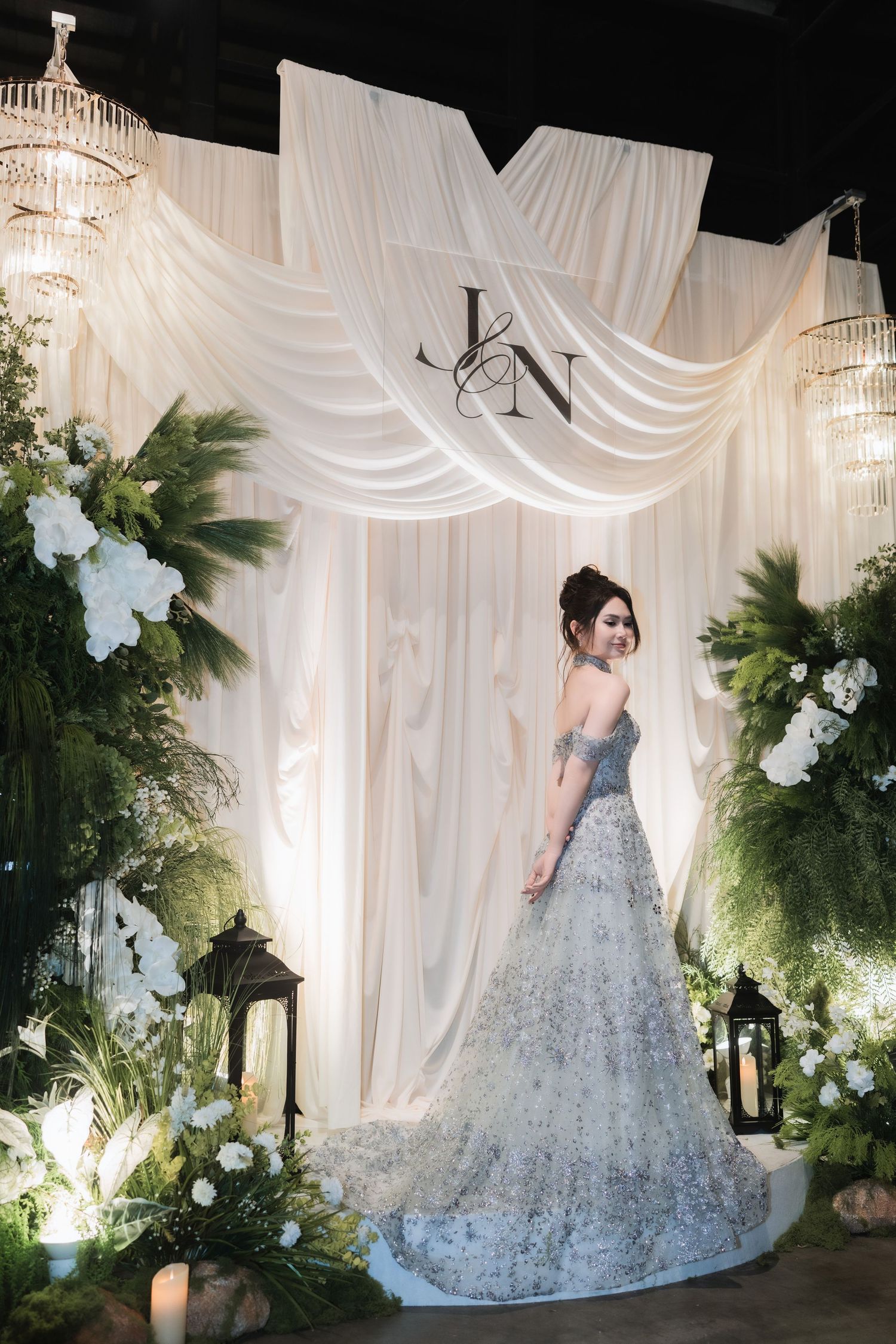 A bride in a sparkling silver gown poses against a white draped backdrop with greenery.