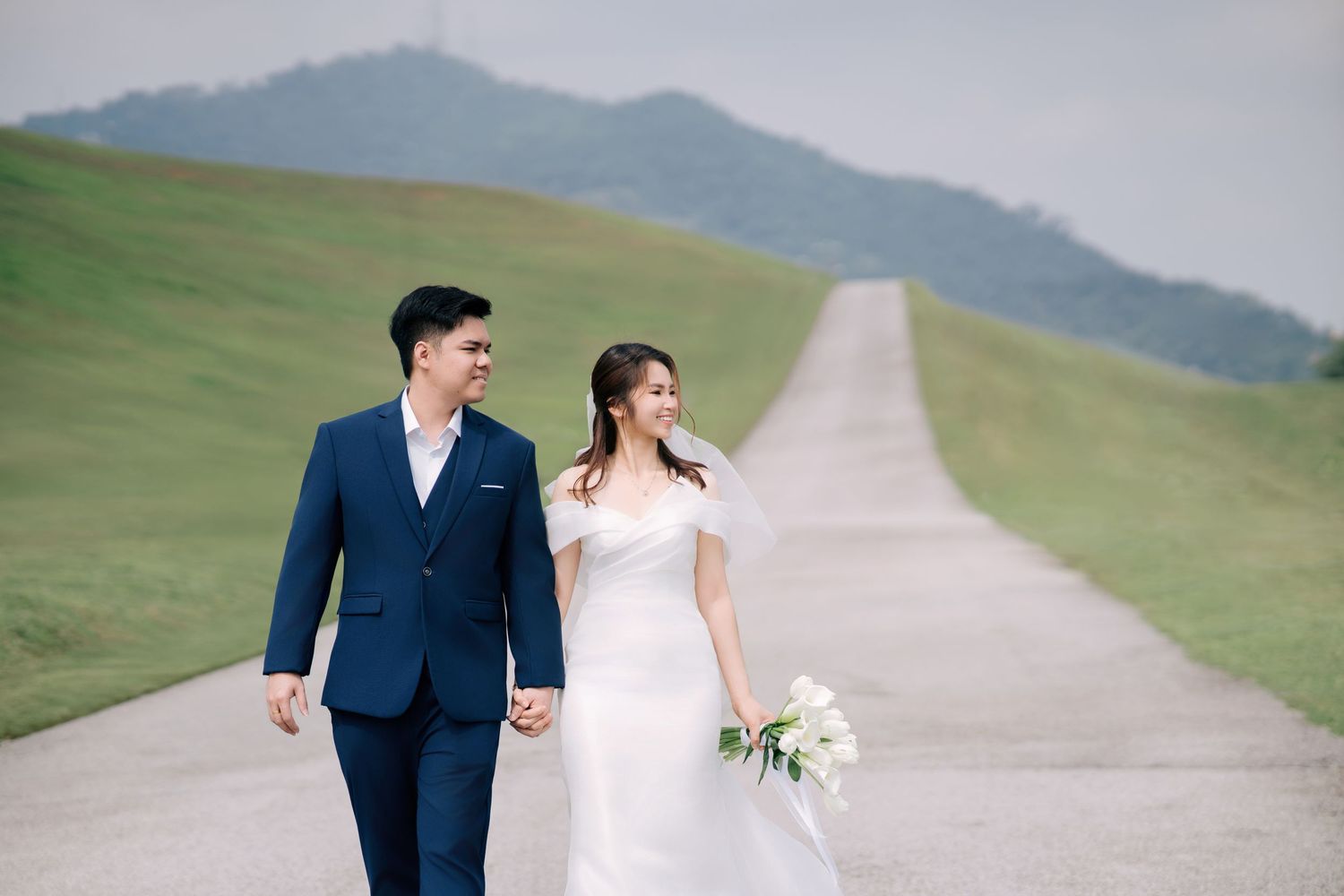 Newlyweds walk hand in hand down a scenic road with mountains in the background.