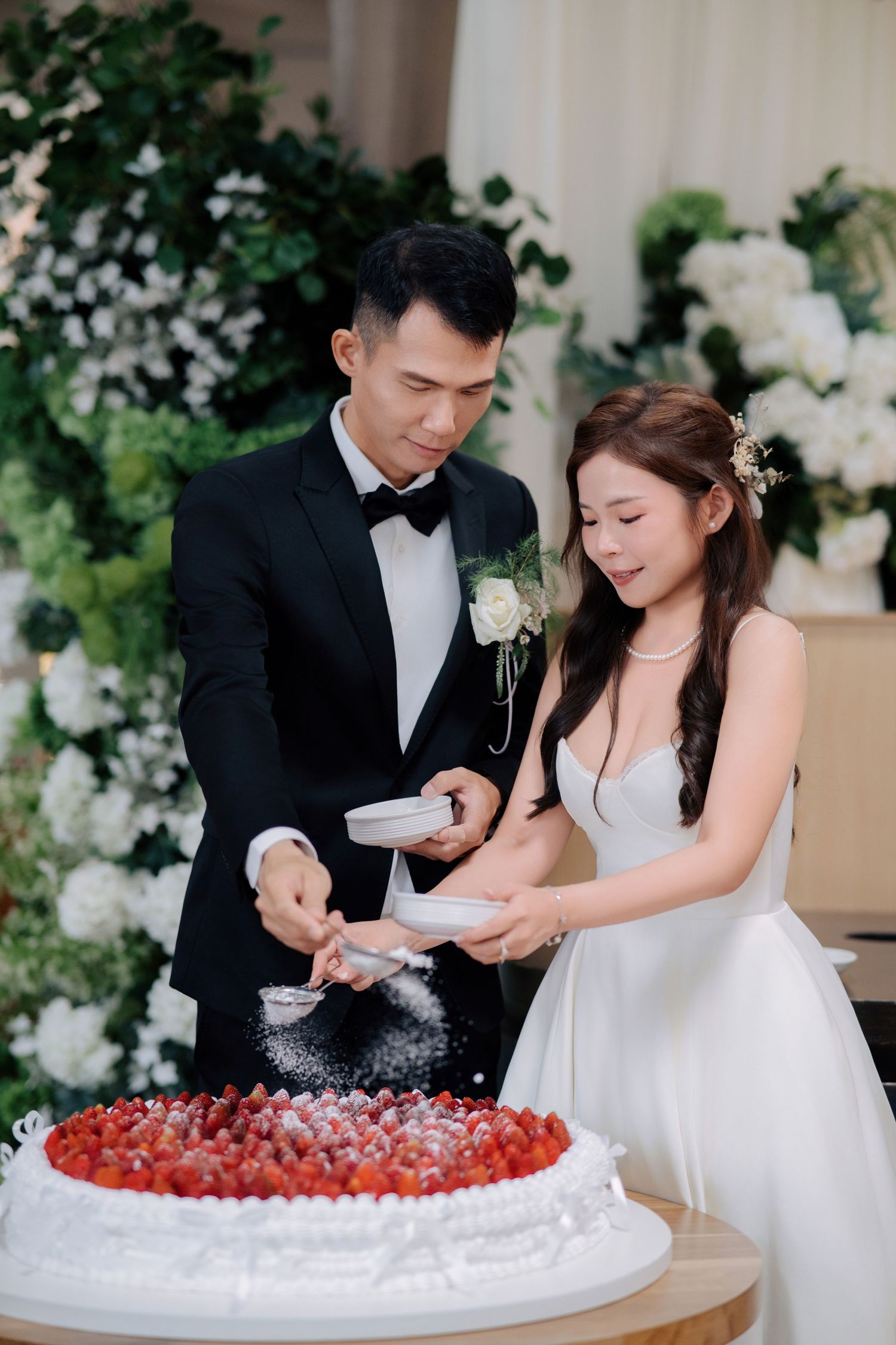 Bride and groom use small sifters to dust powdered sugar over a large, round strawberry cream cake
