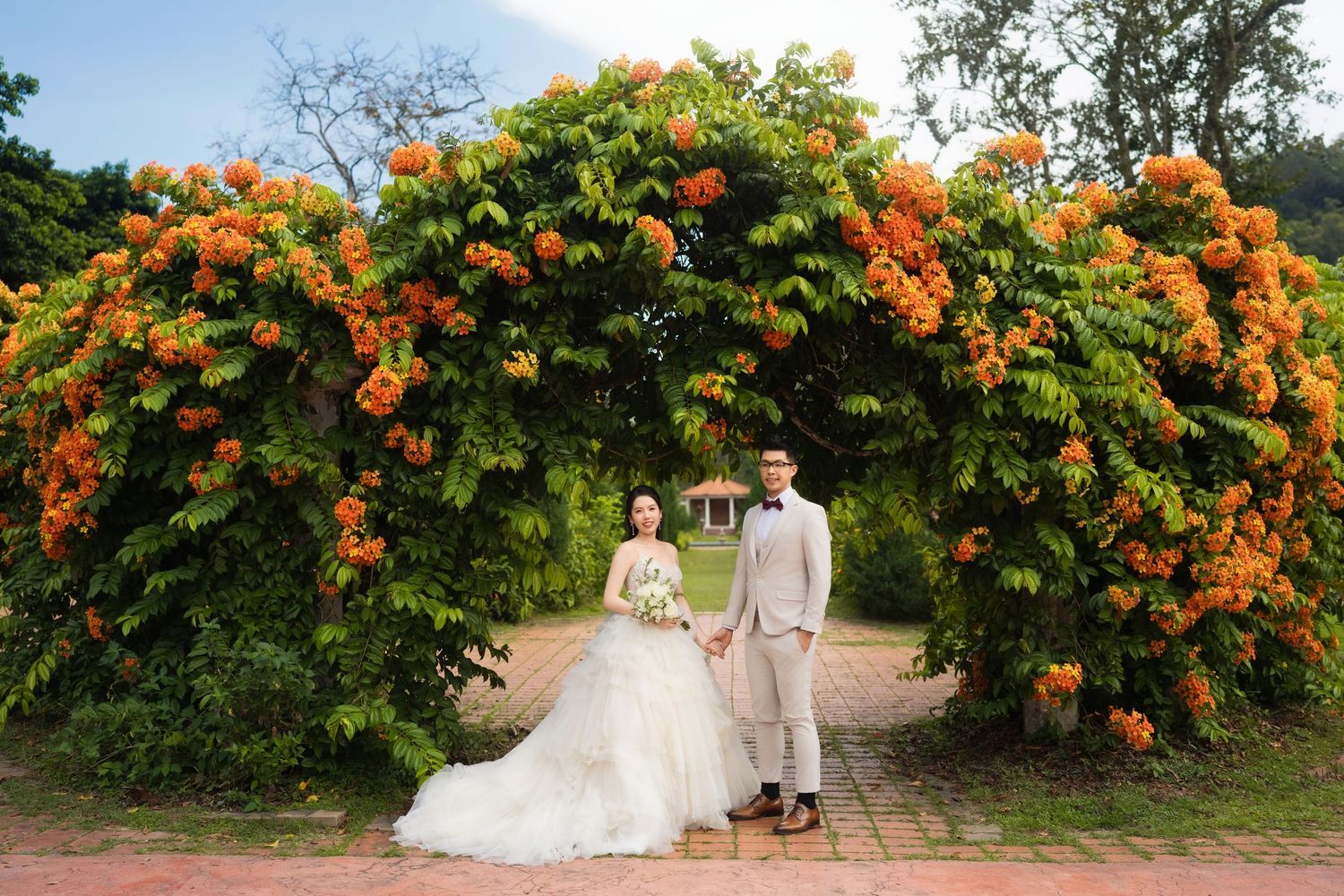 Wedding couple photographed against vibrant orange flowering bushes in garden setting.