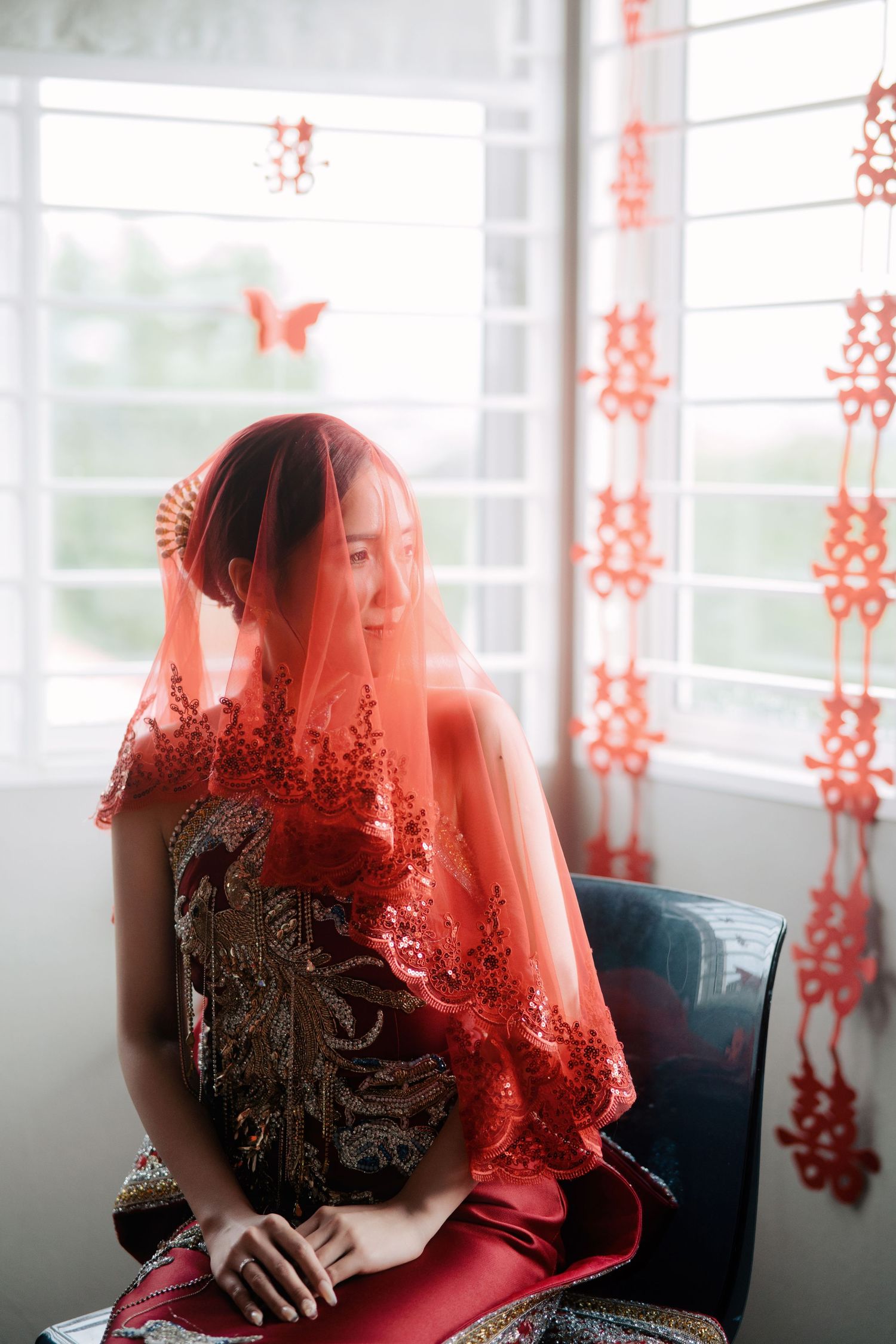 A bride in a red embroidered dress looking to the side through a sheer red lace veil near a window