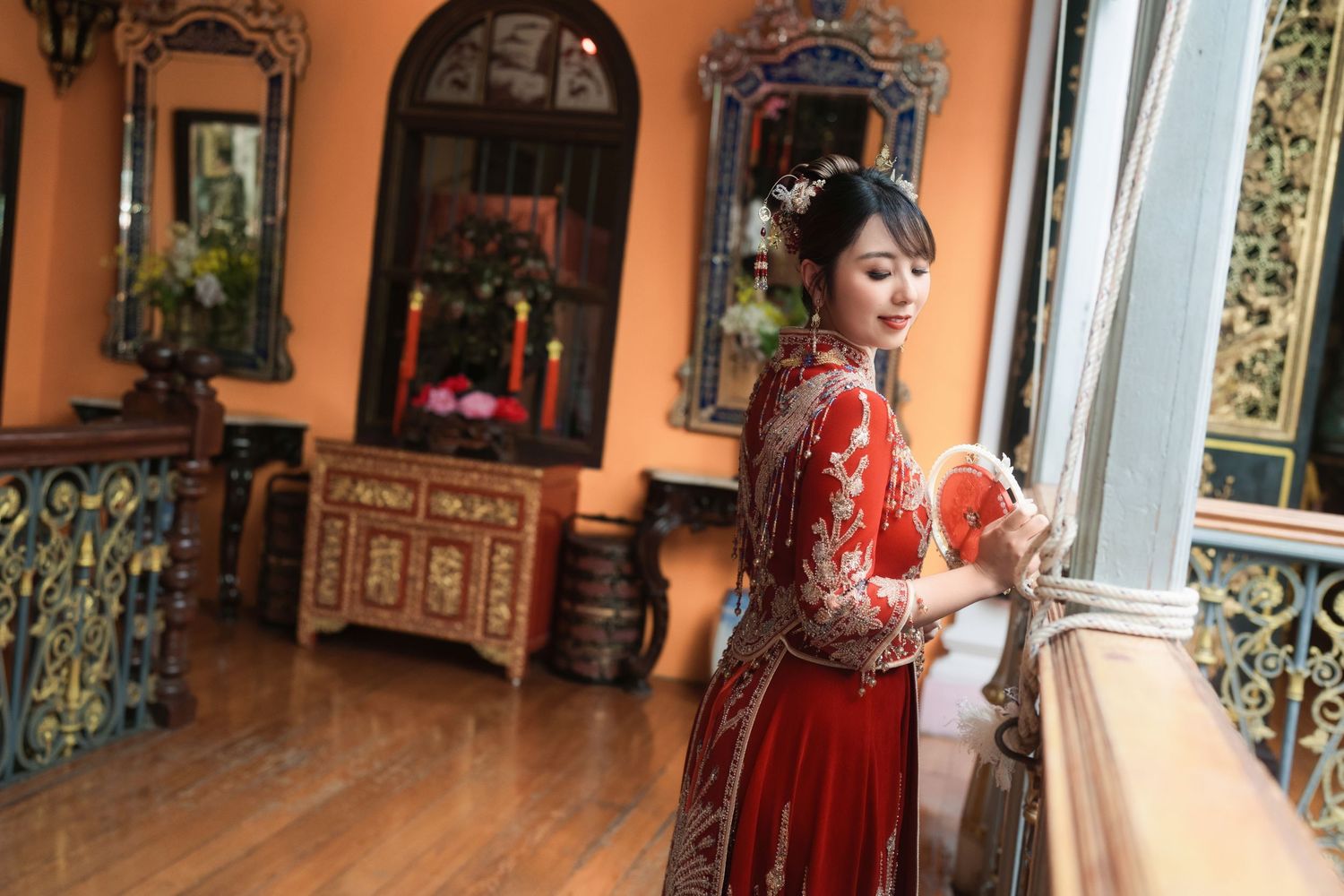 Side view of a bride in a red wedding gown with an intricate updo and gold hair ornaments, looking down at a red fan