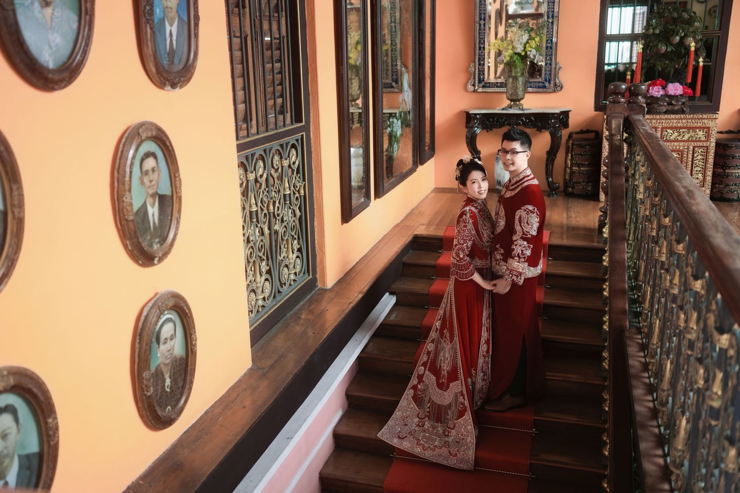 Bride and groom on the red-carpeted staircase of Pinang Peranakan Mansion, surrounded by historical family portraits