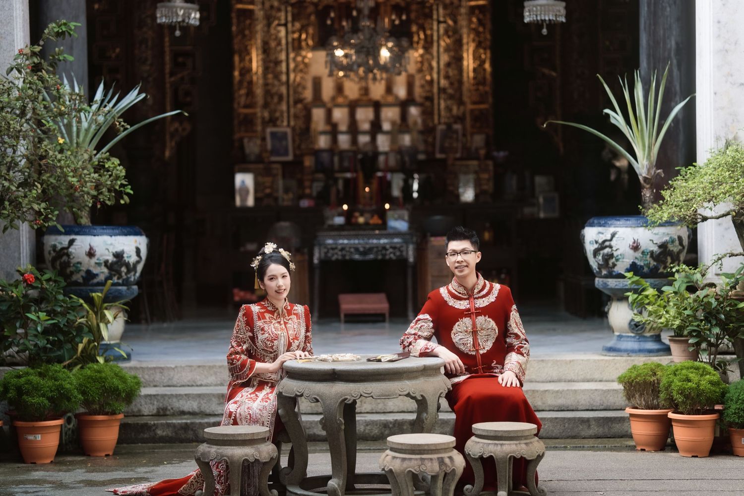 A bride and groom pose in traditional dress at a stone table within the historic courtyard of the Pinang Peranakan Mansion