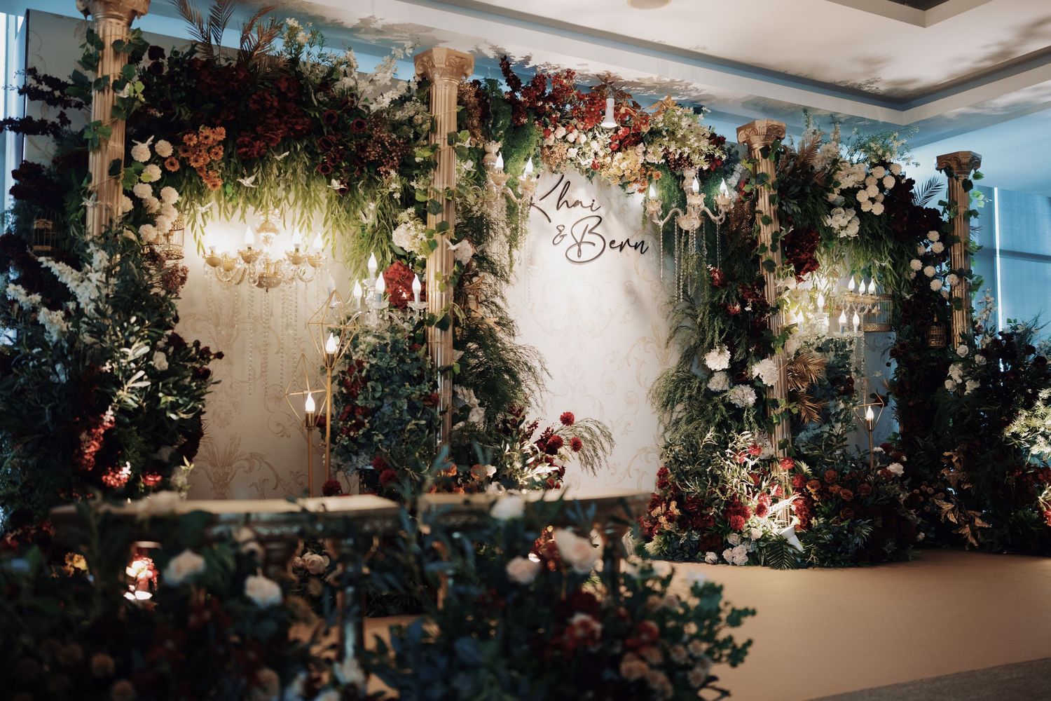 Romantic floral wedding backdrop featuring deep red blooms, gold pillars, and elegant warm chandeliers