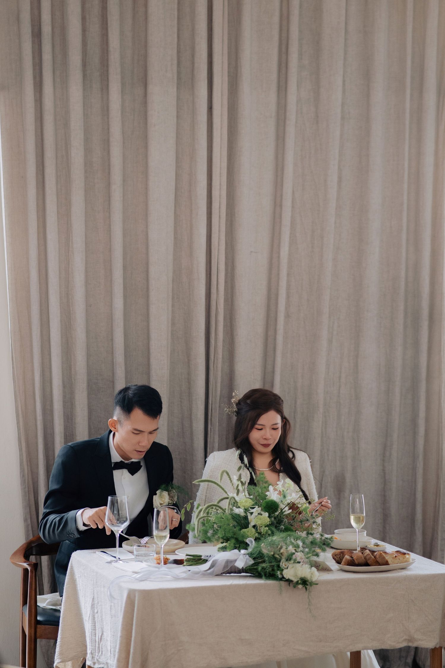 Bride in a white tweed jacket and groom in a tux eat dinner at a candlelit table with green florals