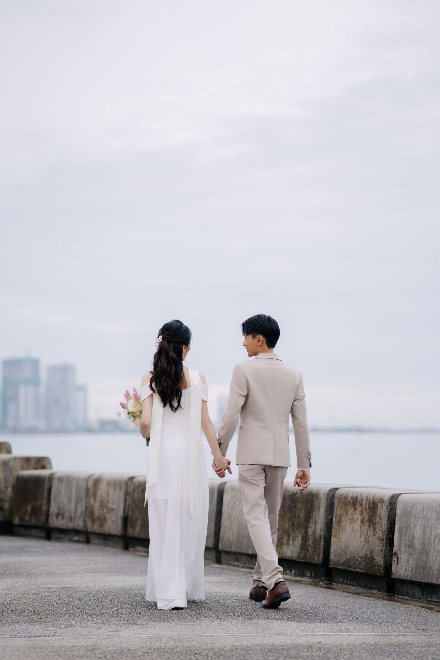 Minimalist wedding photo along concrete seawall with cloudy sky backdrop.