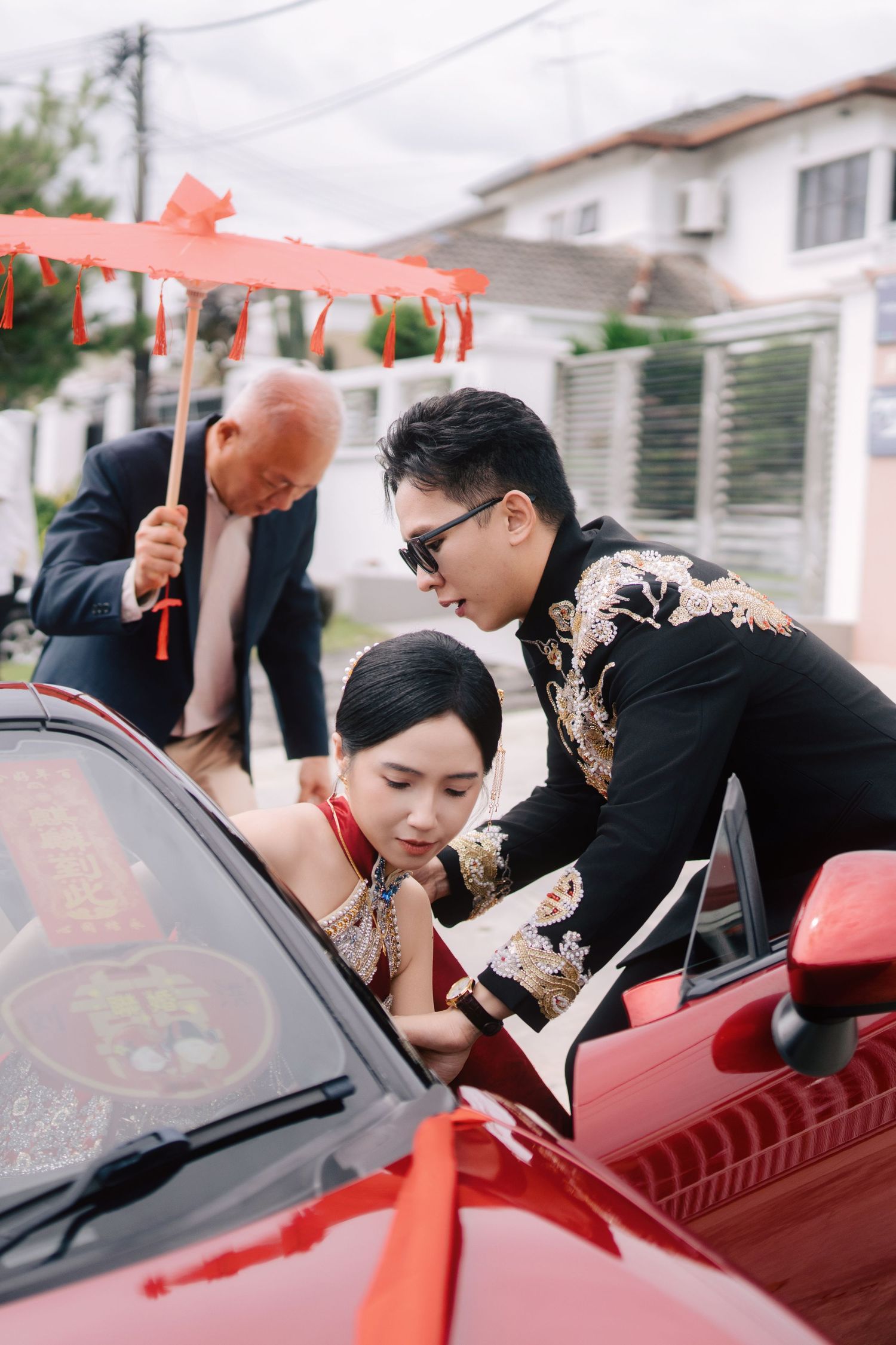 Groom in black Tang suit helping bride in red cheongsam into a red convertible under a red oil-paper umbrella