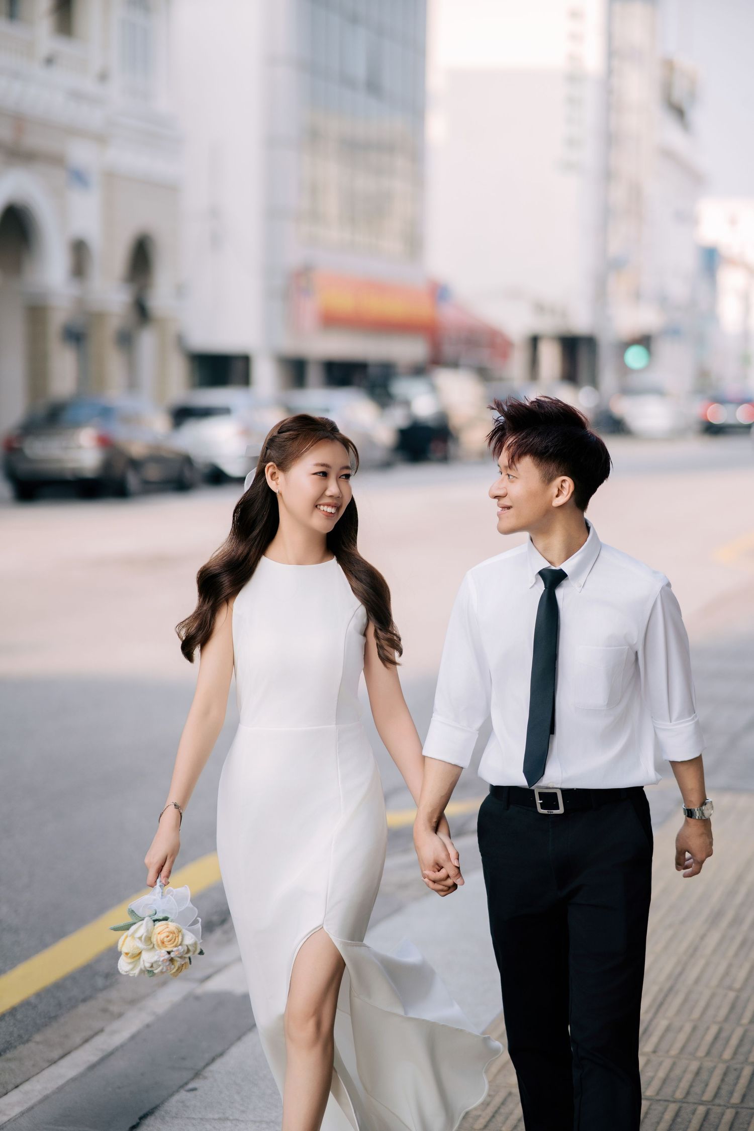 Couple in wedding attire walking hand in hand along a city street.